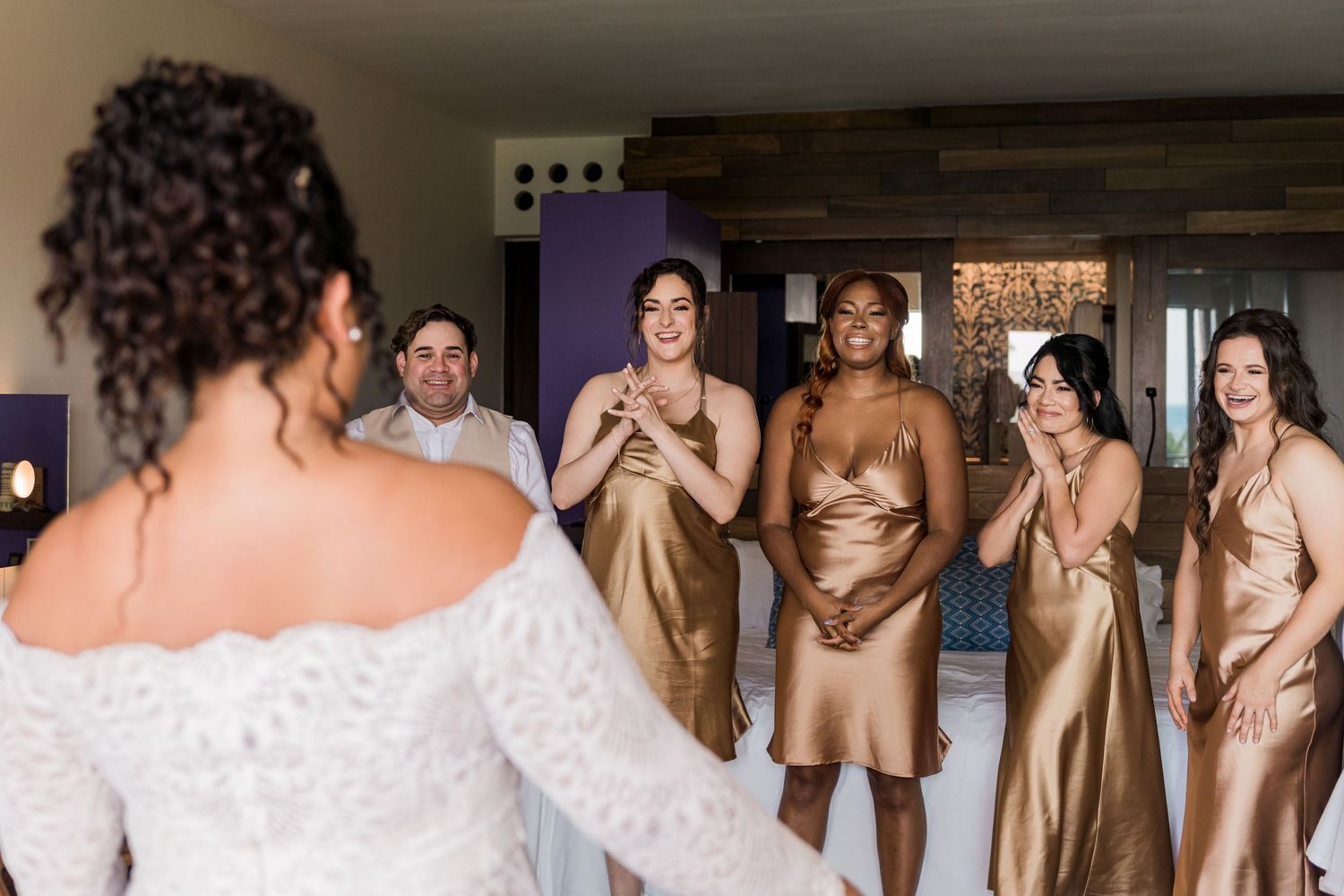 Bride in white lace gown reveals herself to emotional bridesmaids in gold satin dresses during wedding getting ready moment.