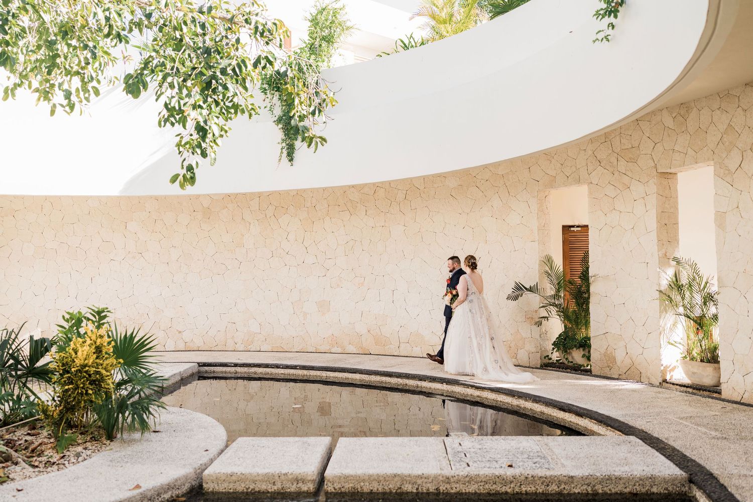 Secrets Akumal Wedding - Bride and groom embrace near a curved white stone wall and reflecting pool in a tropical garden setting.