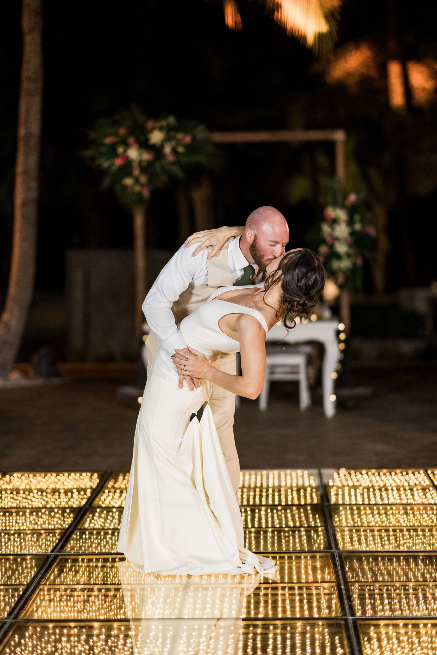Bride and groom share a romantic first dance kiss on a glowing LED dance floor at an outdoor wedding reception at night.