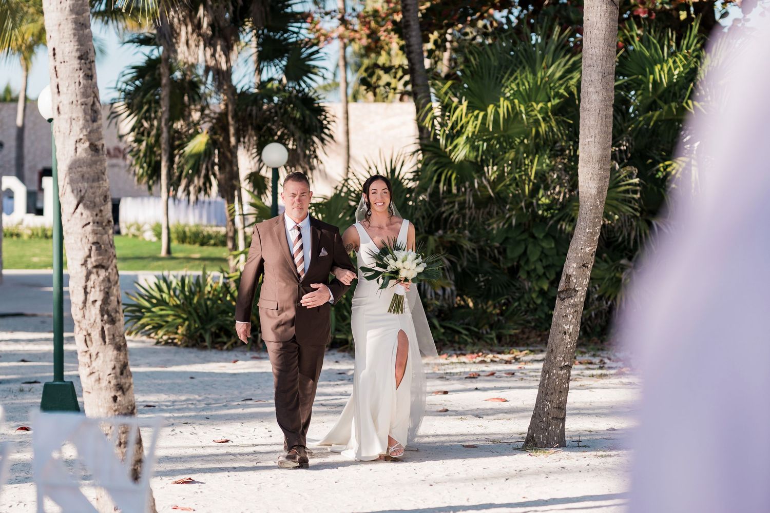 Bride walks down sandy aisle with escort among palm trees at outdoor beach wedding ceremony in tropical setting.