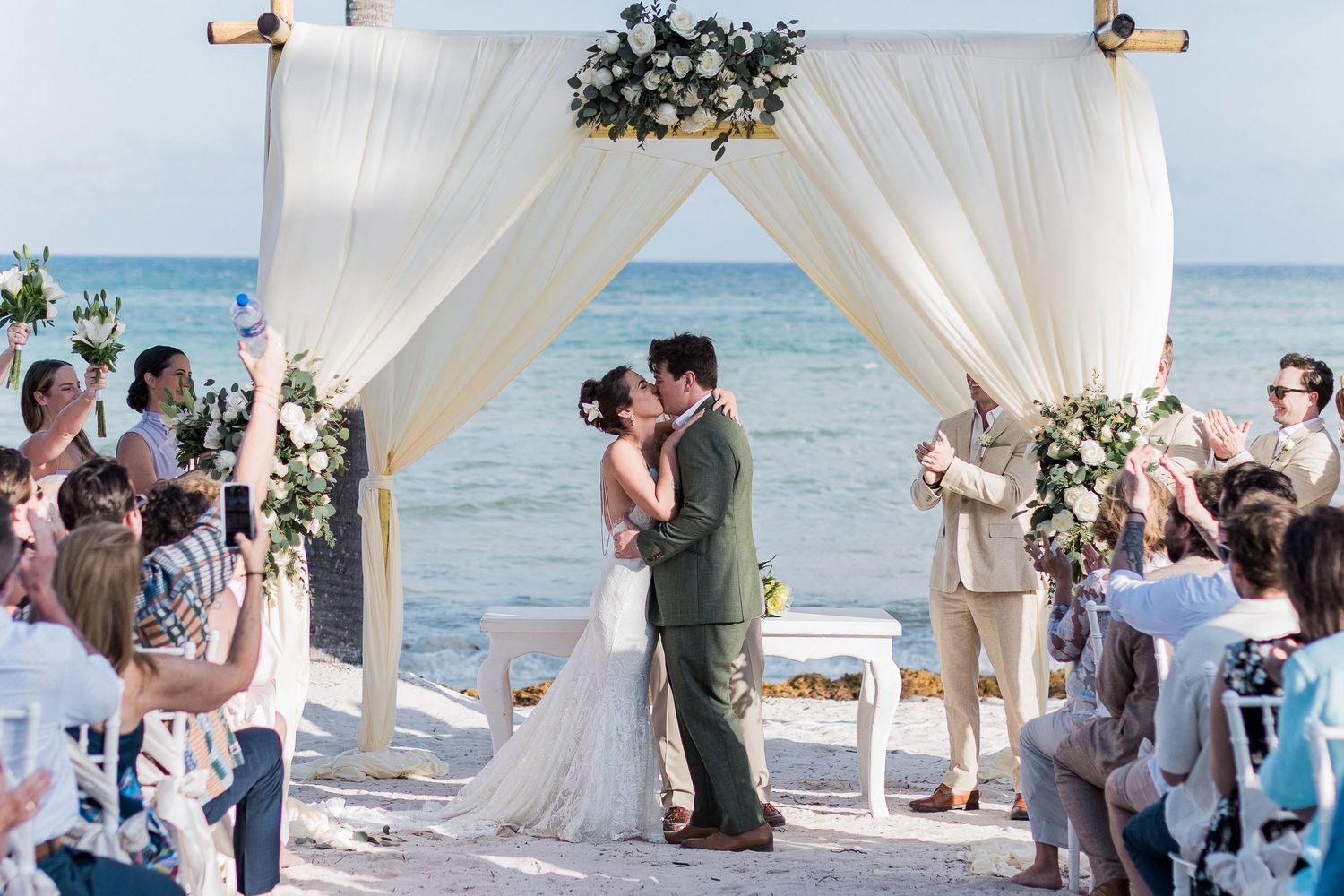 Couple shares first kiss at romantic beach wedding ceremony under floral arch with ocean backdrop.
