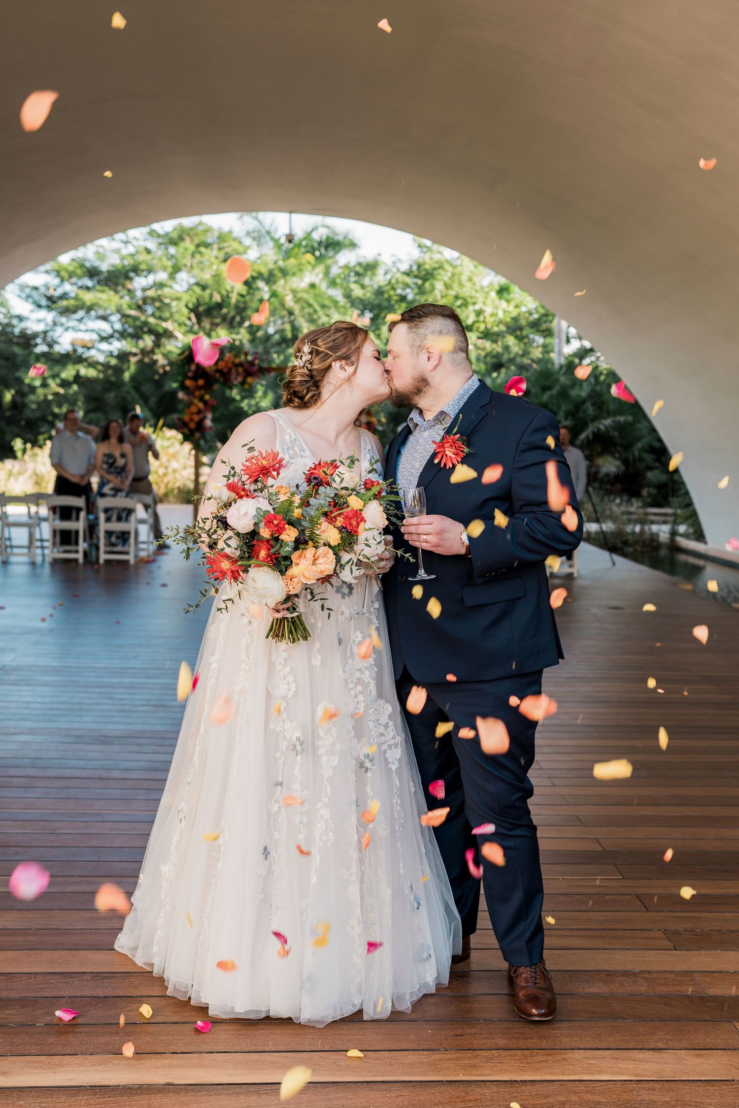 Secrets Akumal Wedding - Bride and groom kissing under arch with colorful flower petals falling around them on wooden dance floor.