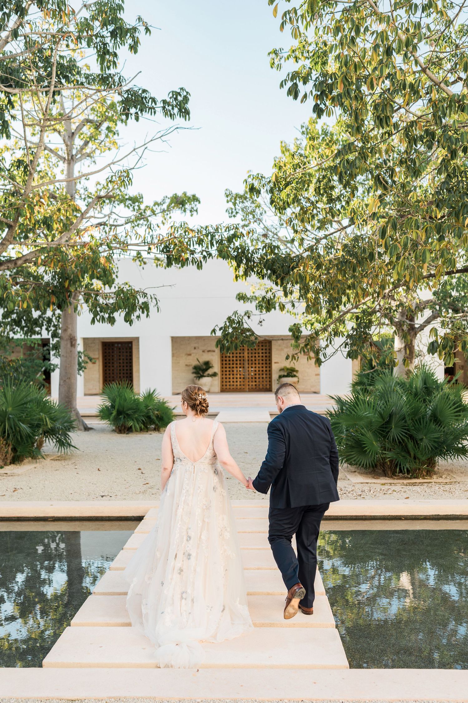Secrets Akumal Wedding - Bride and groom walk hand in hand toward a reflecting pool surrounded by lush trees at an elegant outdoor venue.