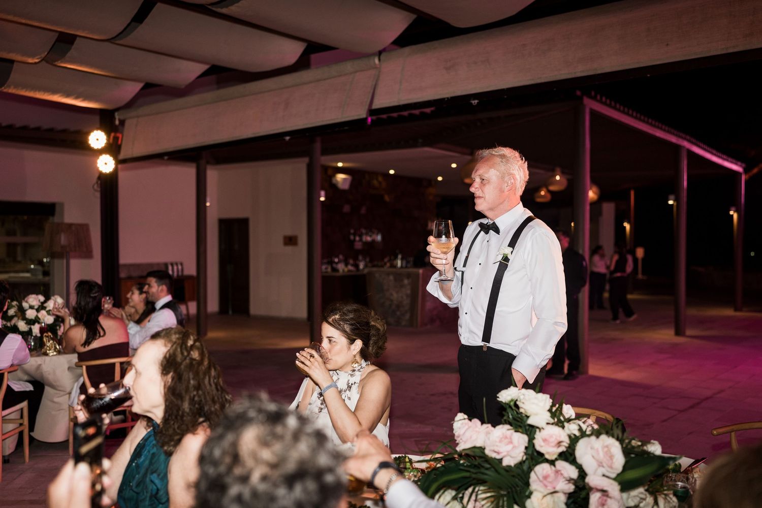 Man in white shirt and suspenders giving a toast at an elegant wedding reception with floral arrangements.