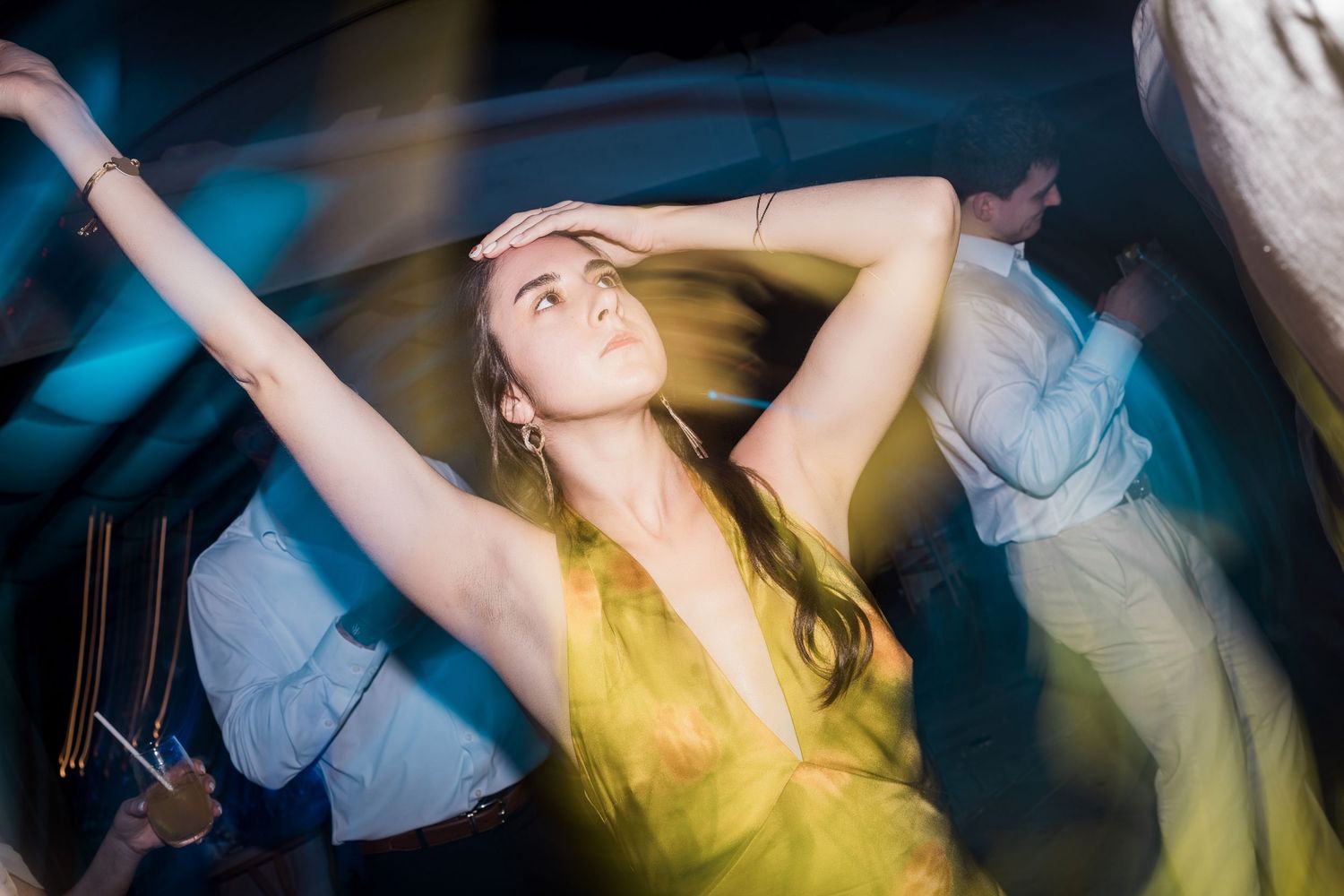 Woman in yellow dress dancing at a party, arms raised, under blue and yellow lights.