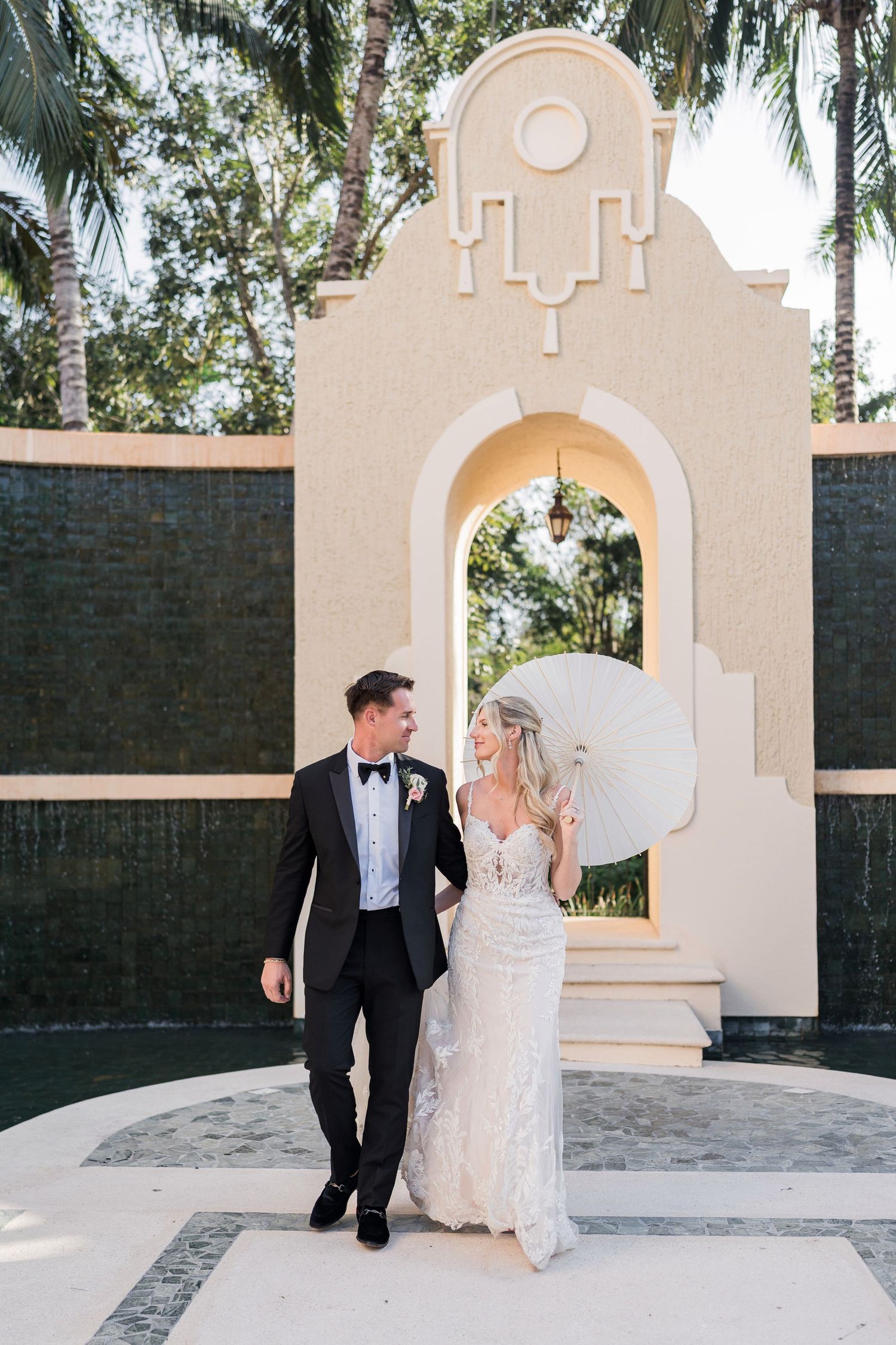 Bride in lace gown and groom in black tuxedo walking together in front of elegant arch at tropical wedding venue.