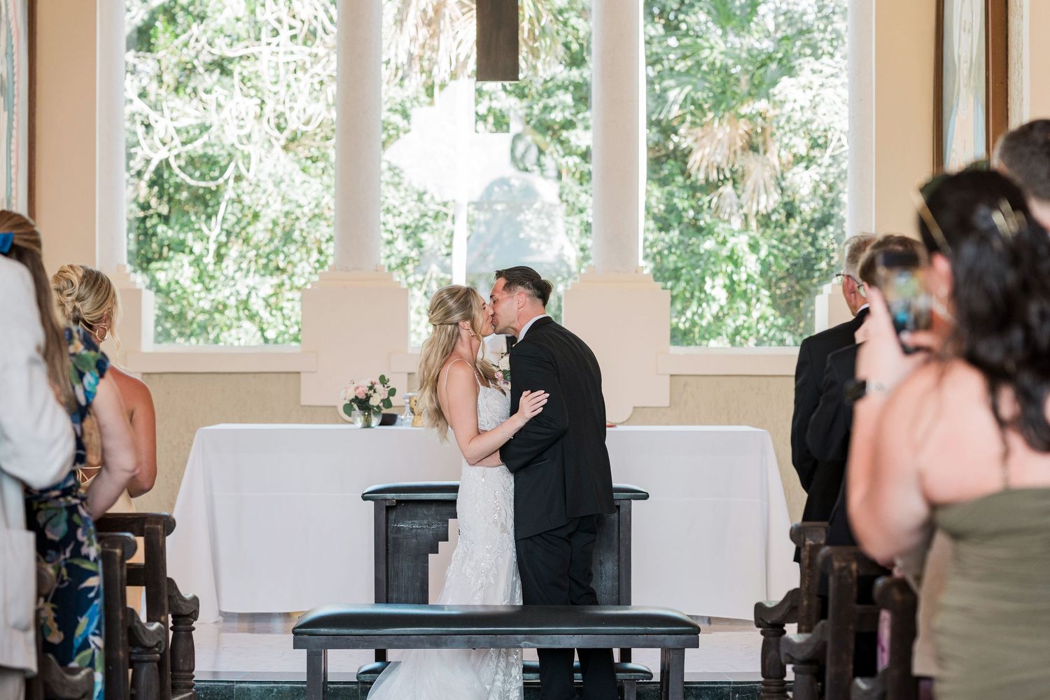 Bride and groom share first kiss at wedding altar in elegant white chapel with large windows.