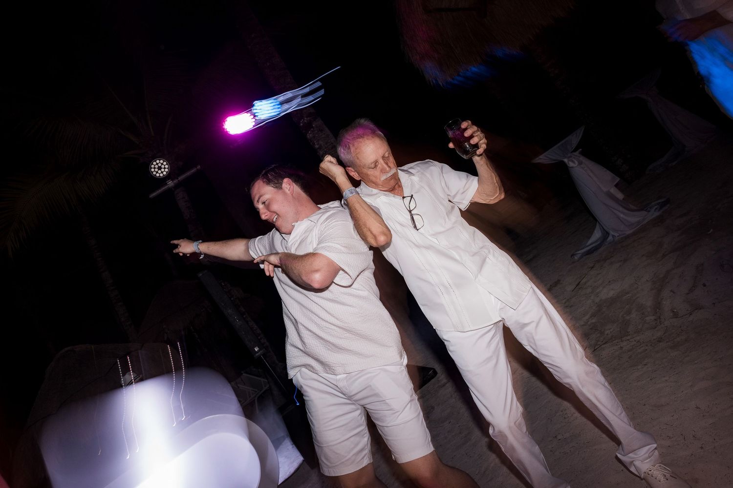 Two people in white outfits dancing energetically at a nighttime outdoor party under colorful lights.