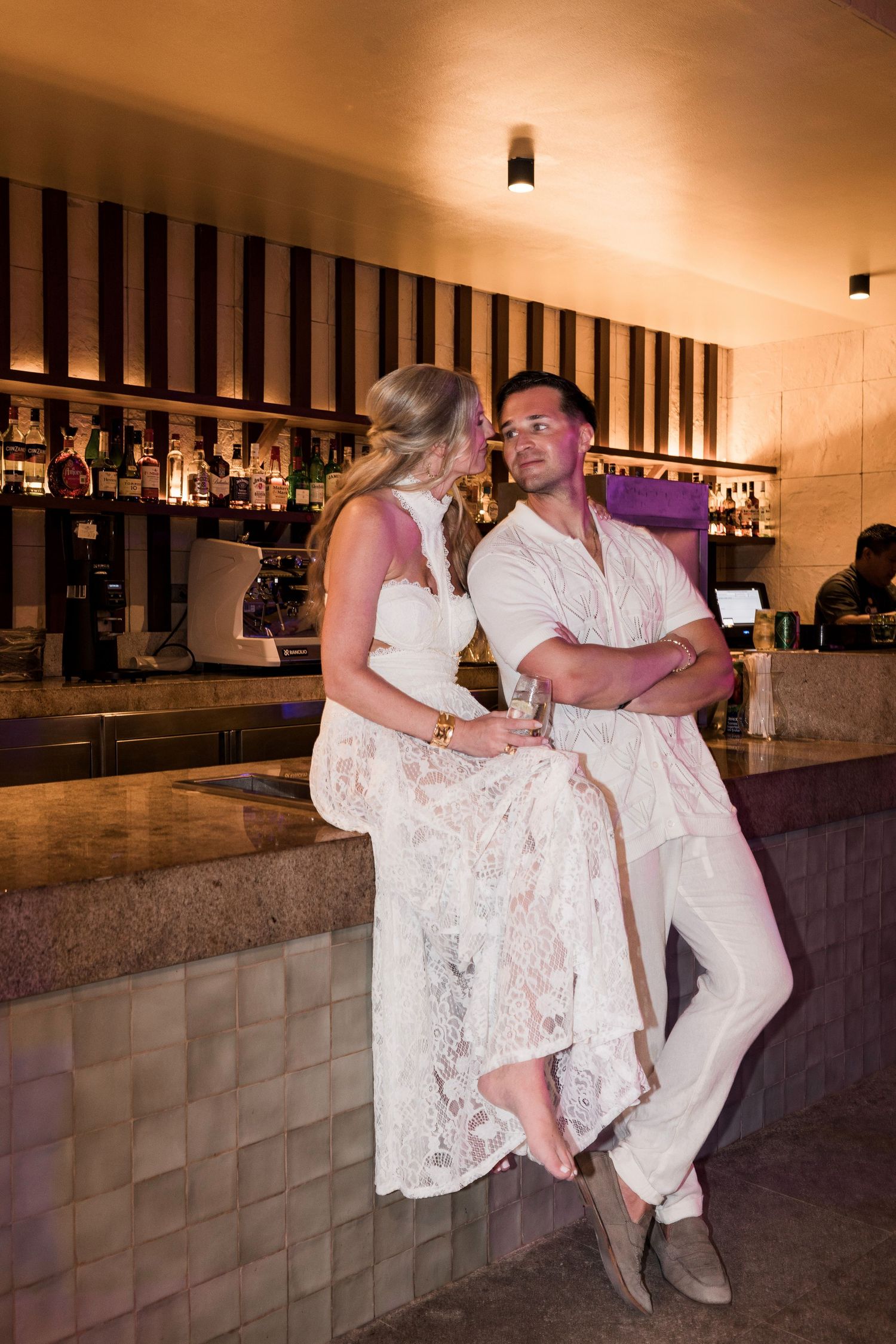 Couple in white outfits posing together at a bar, woman sitting on counter, man leaning beside her.
