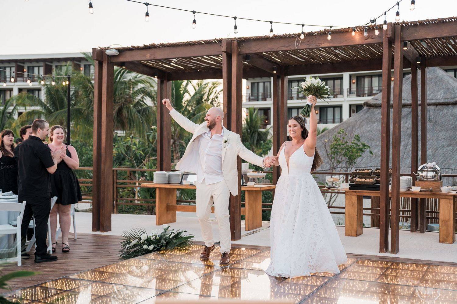 Bride and groom make grand entrance at outdoor wedding reception with string lights and tropical pergola backdrop.
