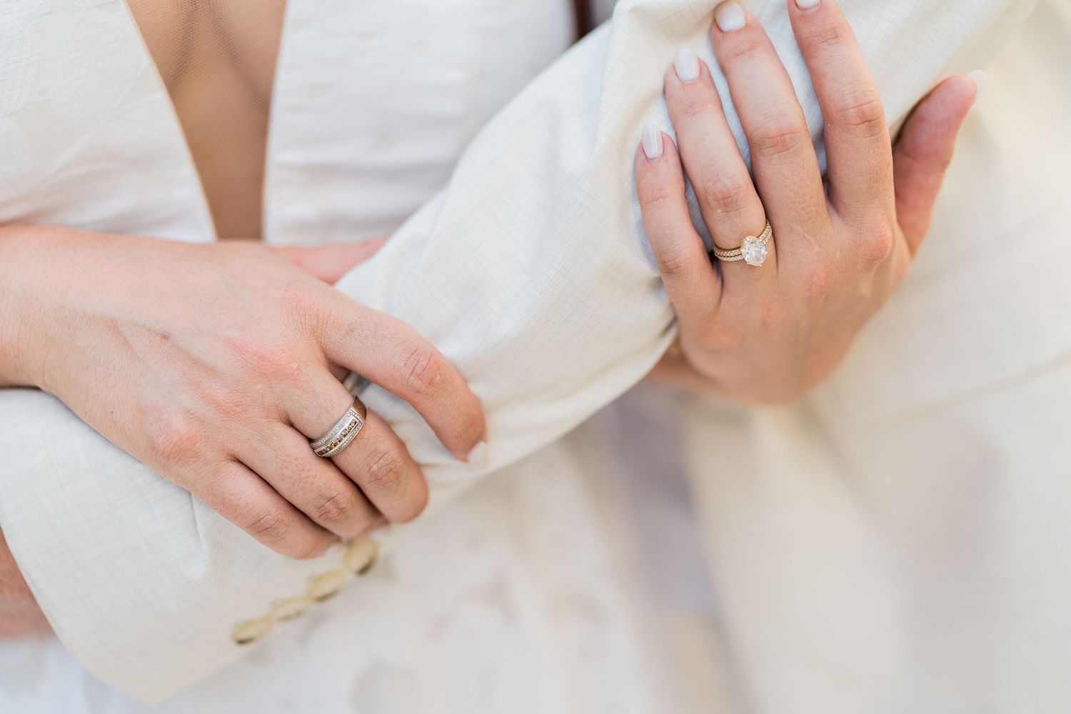 Two brides wearing wedding rings hold hands, showcasing elegant engagement and wedding bands on their fingers.
