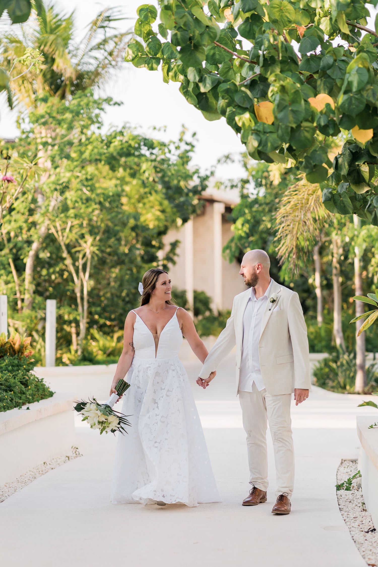 Bride and groom hold hands walking on a tropical path, she in lace gown with bouquet, he in tan suit.