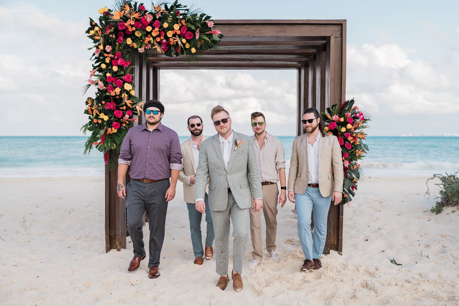 Five groomsmen posing on a beach wedding altar decorated with tropical flowers, with ocean waves in the background.