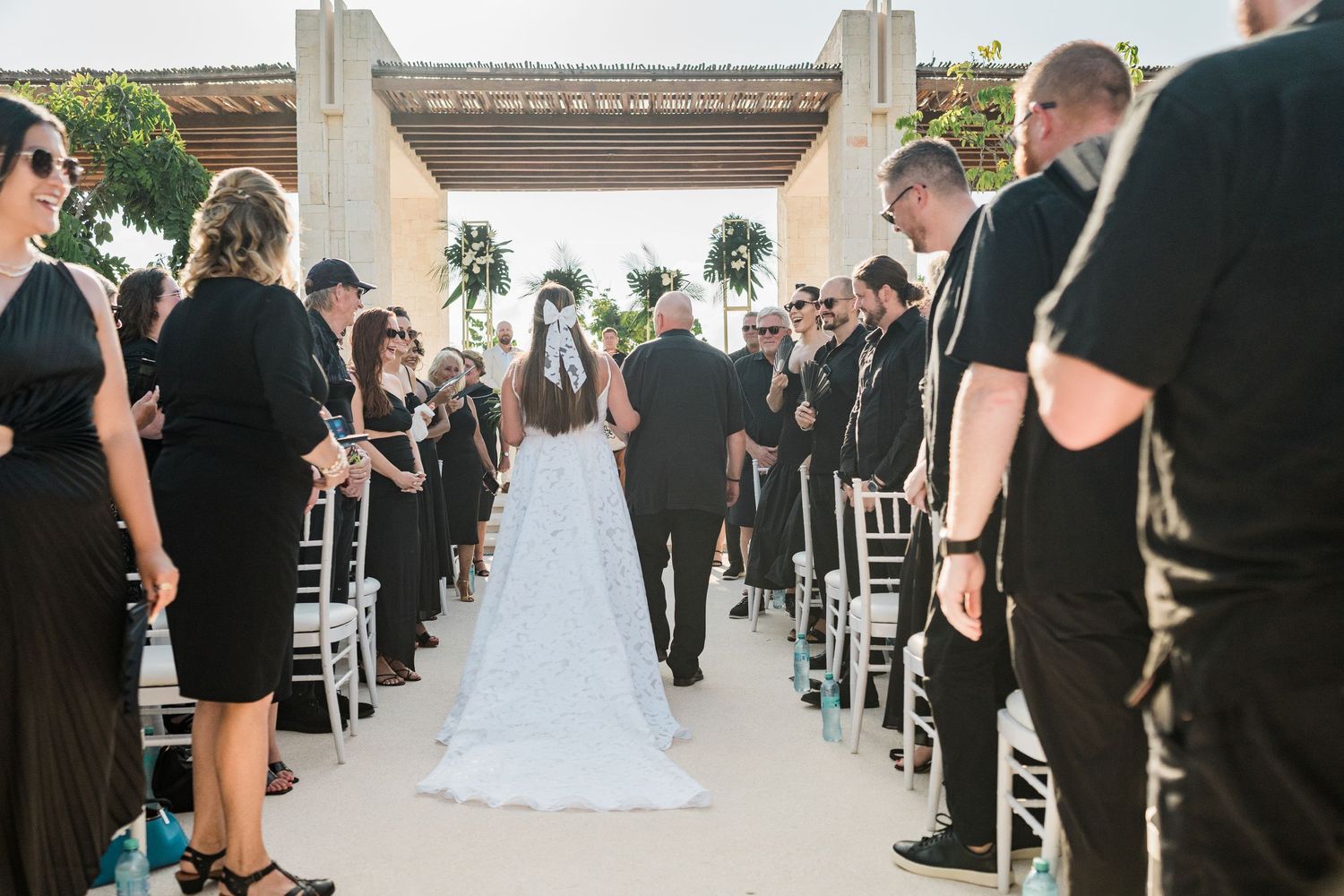 Bride walks down the aisle at outdoor wedding ceremony with guests dressed in black lining the path.
