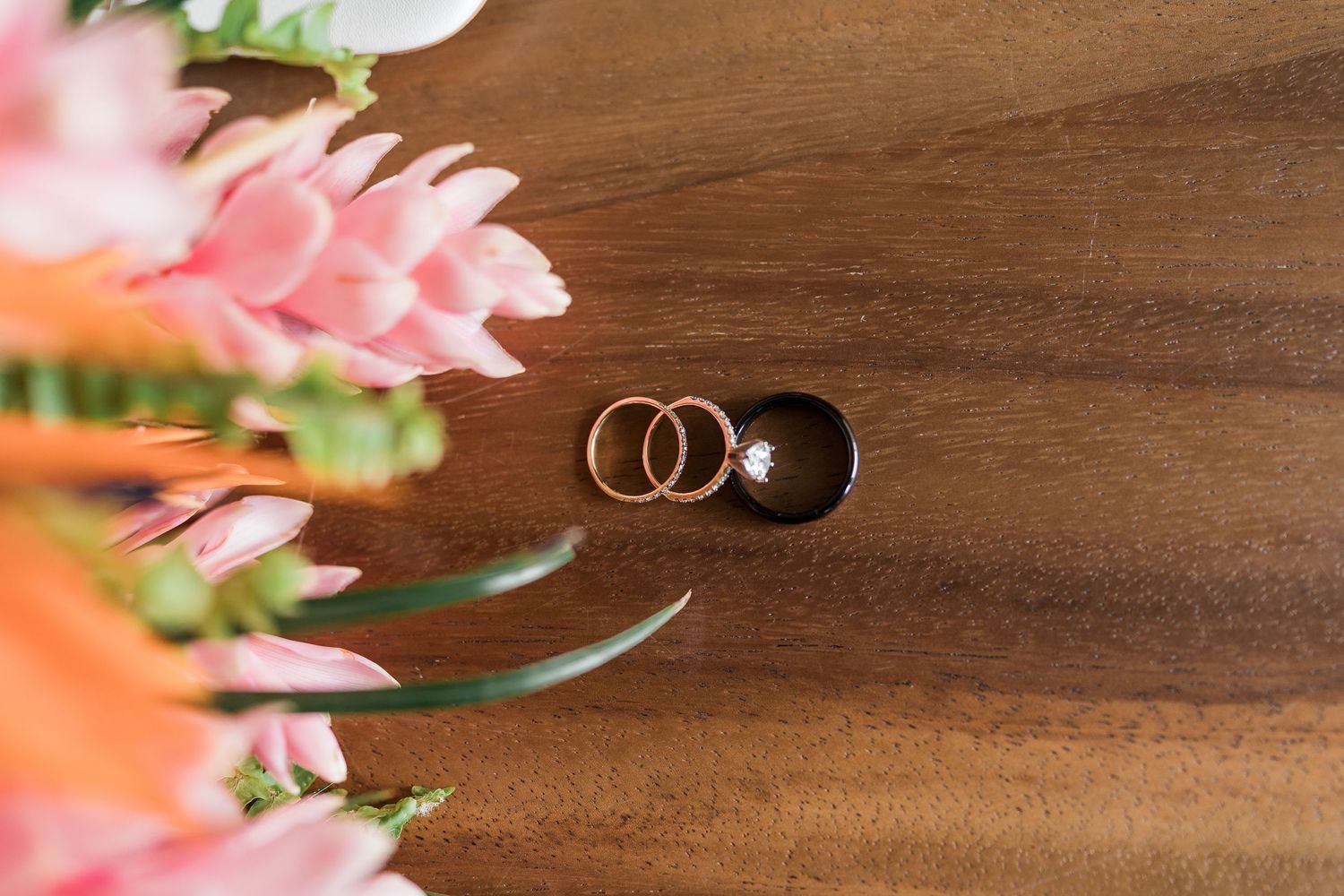 Two gold wedding rings and a diamond engagement ring on a wooden surface with pink flowers in the background.