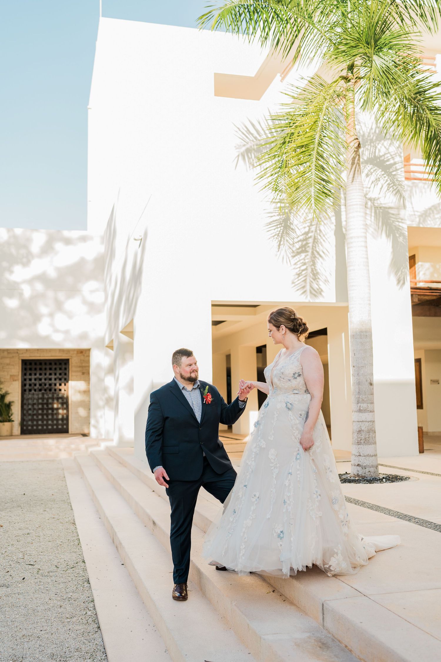 Bride and groom holding hands on steps outside a white building with palm trees at a destination wedding at Secrets akumal