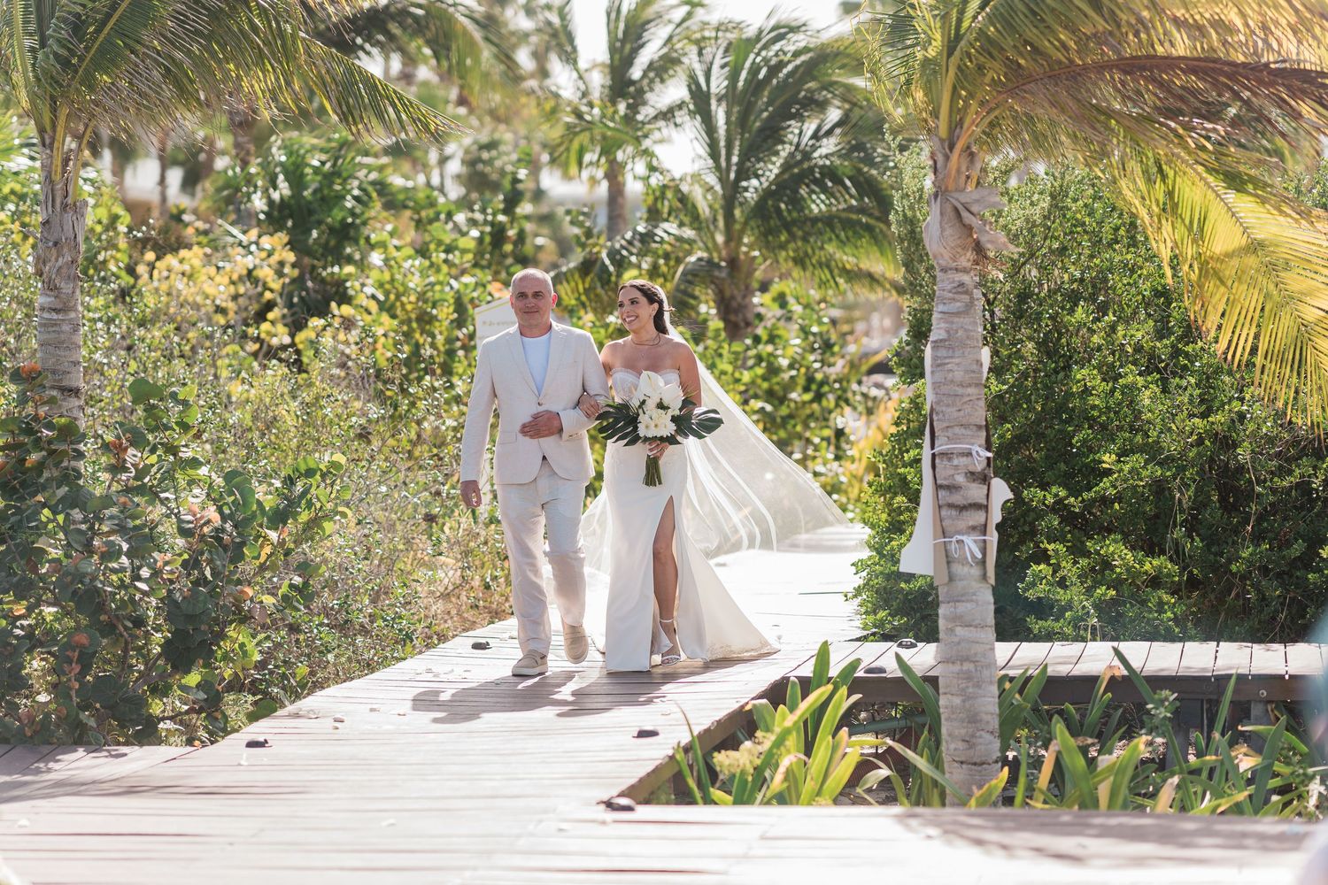 Bride and groom walking hand in hand on a wooden boardwalk surrounded by tropical palms at a beach wedding.