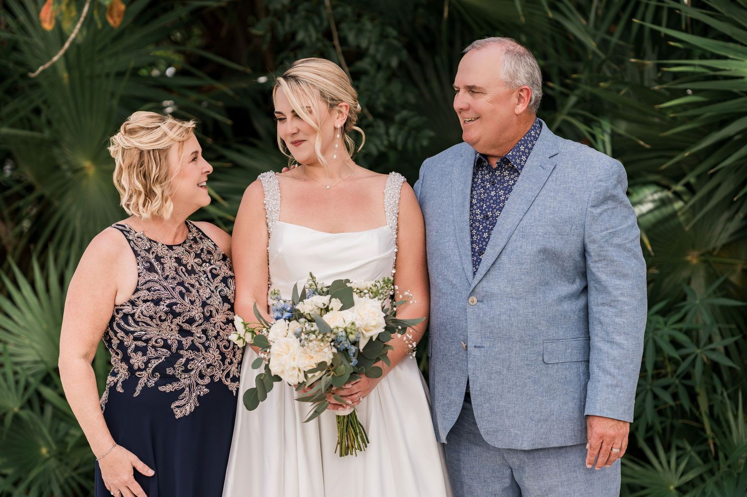 Bride in white gown holds bouquet flanked by smiling woman in navy dress and man in gray suit outdoors.