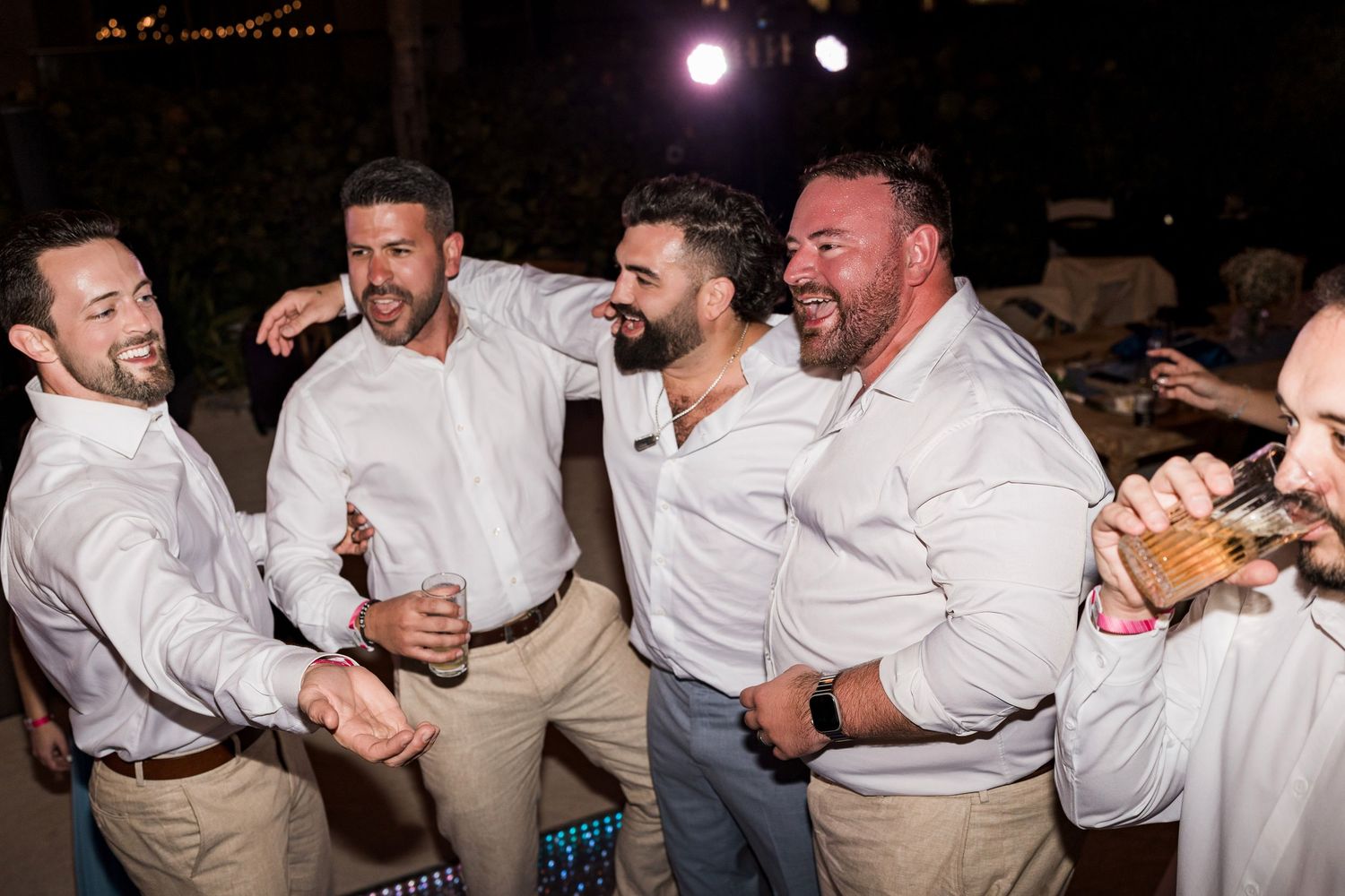 Group of groomsmen laughing and celebrating together at an outdoor wedding reception at night.