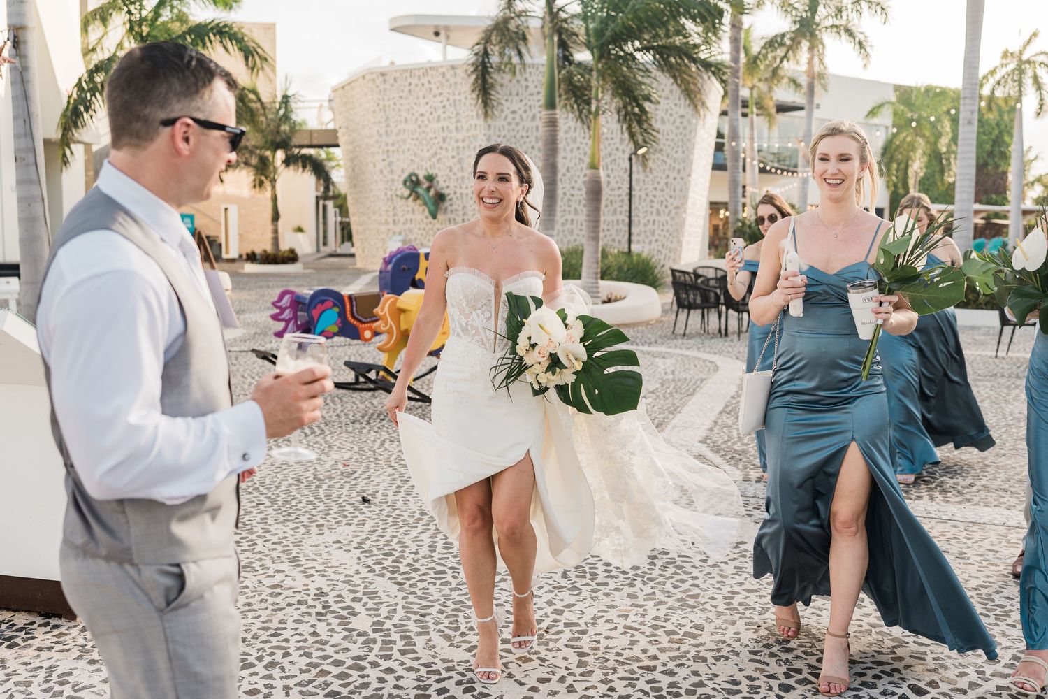 Bride in white dress walks with bridesmaid in blue gown toward groom at tropical outdoor wedding venue.