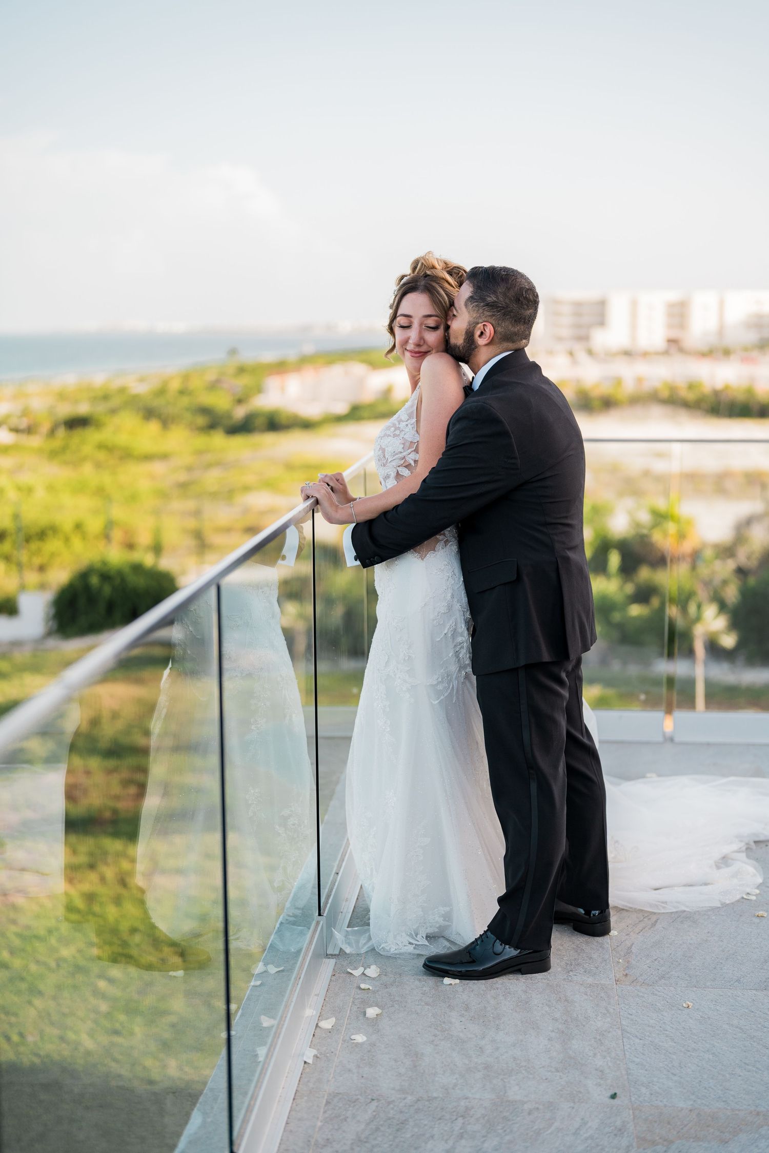 Groom tenderly kisses bride on cheek as they embrace on glass balcony with ocean view.