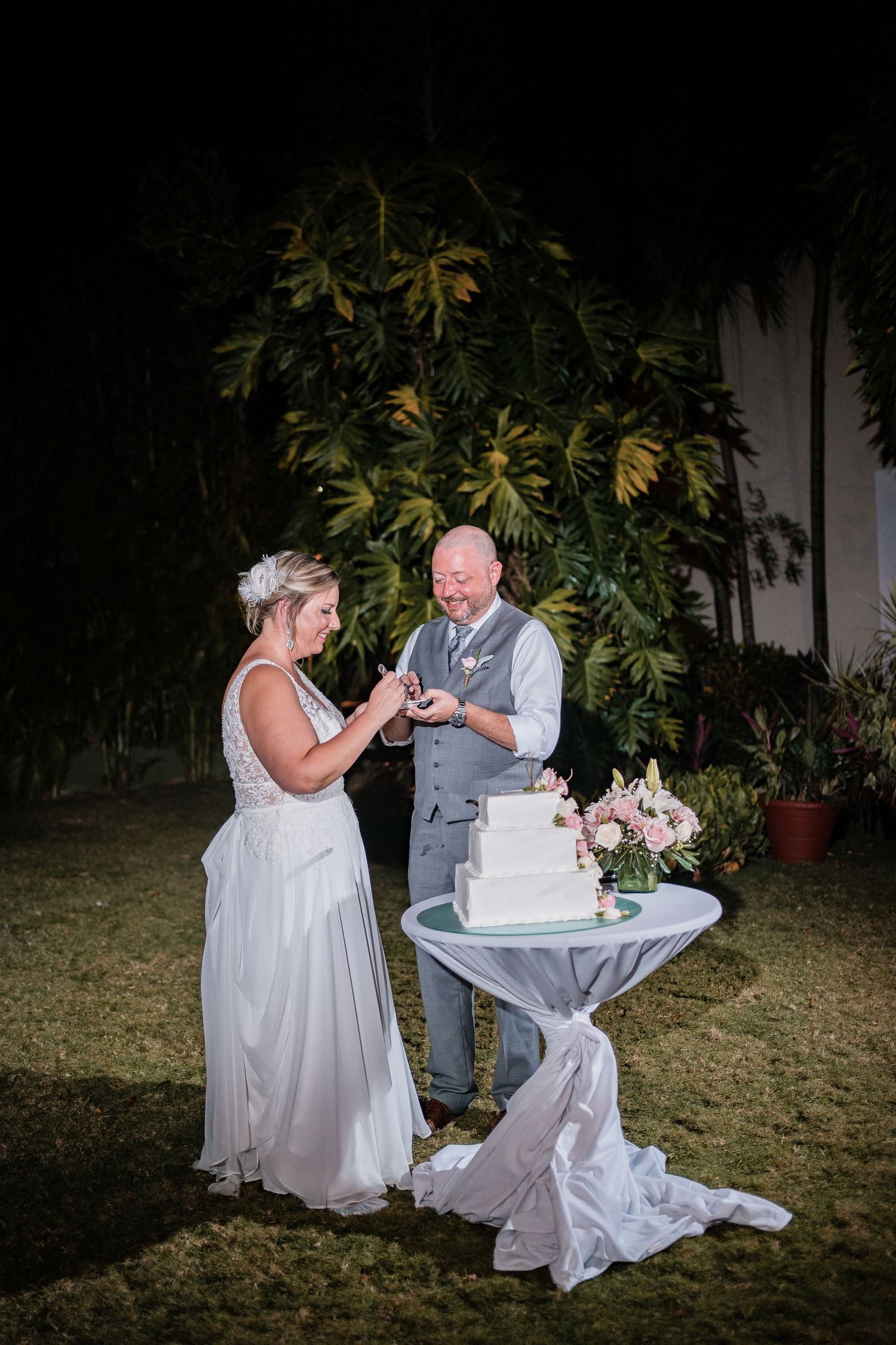 Bride and groom cutting their white tiered wedding cake at an outdoor evening garden reception.