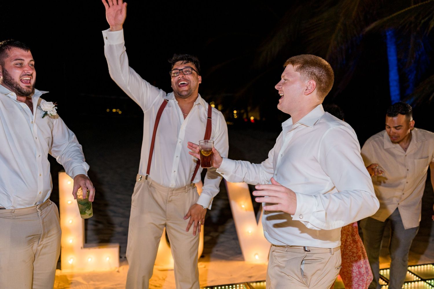 Groomsmen in white shirts and suspenders celebrating near LOVE marquee letters at wedding.