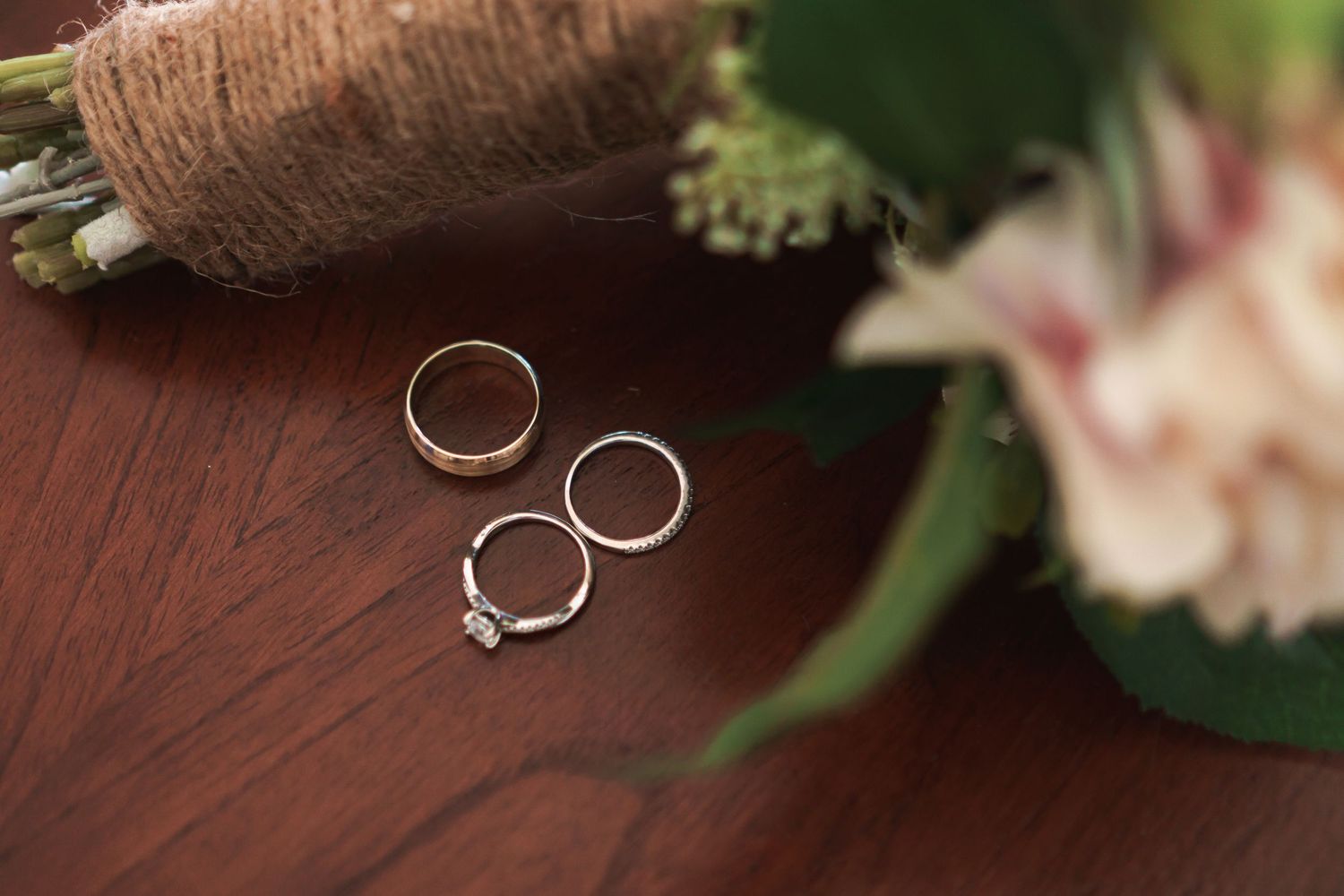 Three wedding rings arranged on wooden surface beside pink and white floral bouquet.