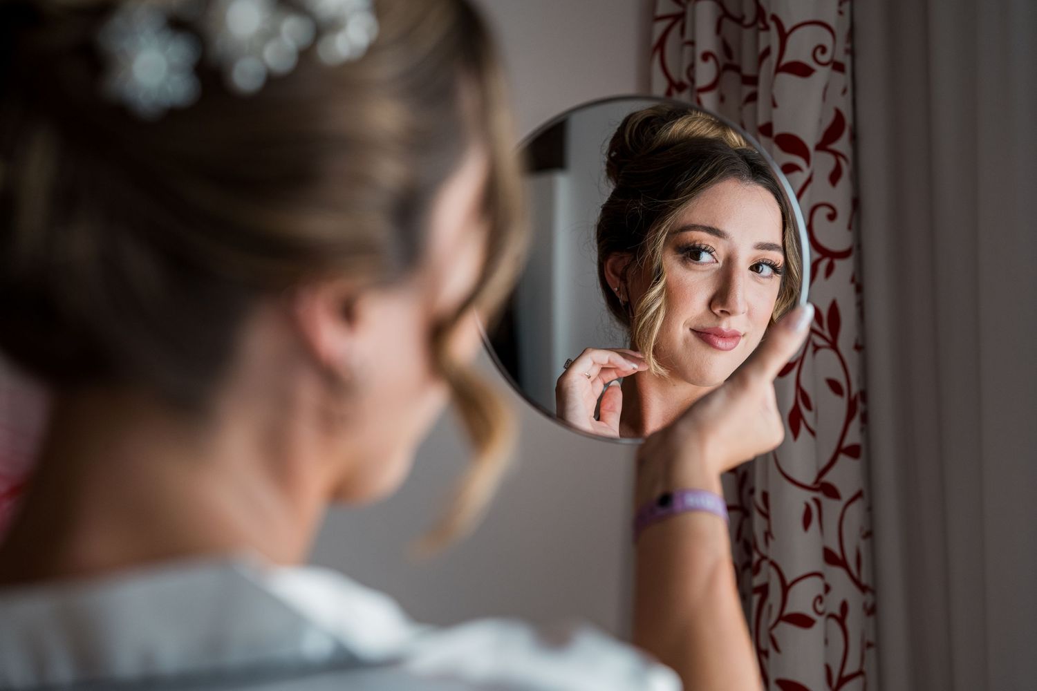 Bride with updo hairstyle checking her makeup in a handheld mirror before her wedding ceremony.