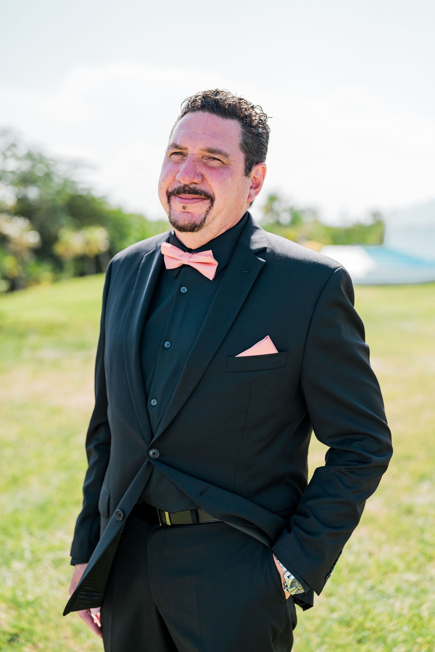 Groom in black tuxedo with pink bow tie and pocket square outdoors.