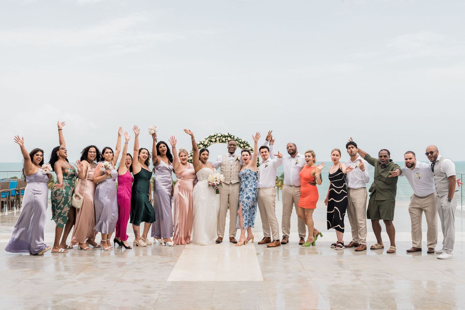 Wedding party group celebrates with raised arms on beachfront terrace with floral arch.