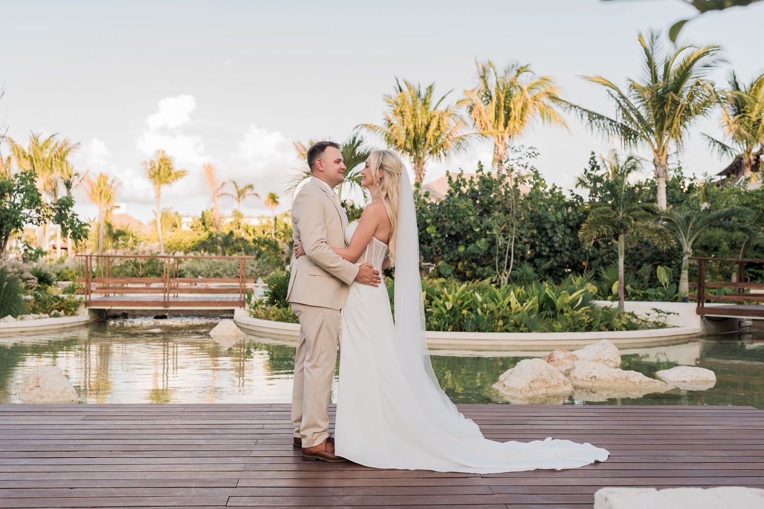 Bride and groom share a romantic moment near a tranquil pond surrounded by palm trees at sunset.