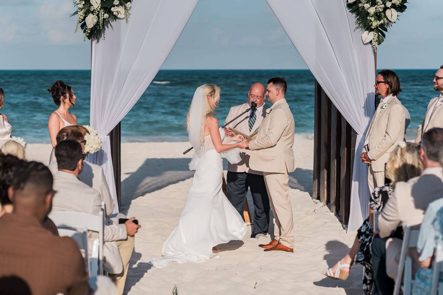 Bride and groom exchange vows under draped floral arch at beach wedding with ocean waves behind them.