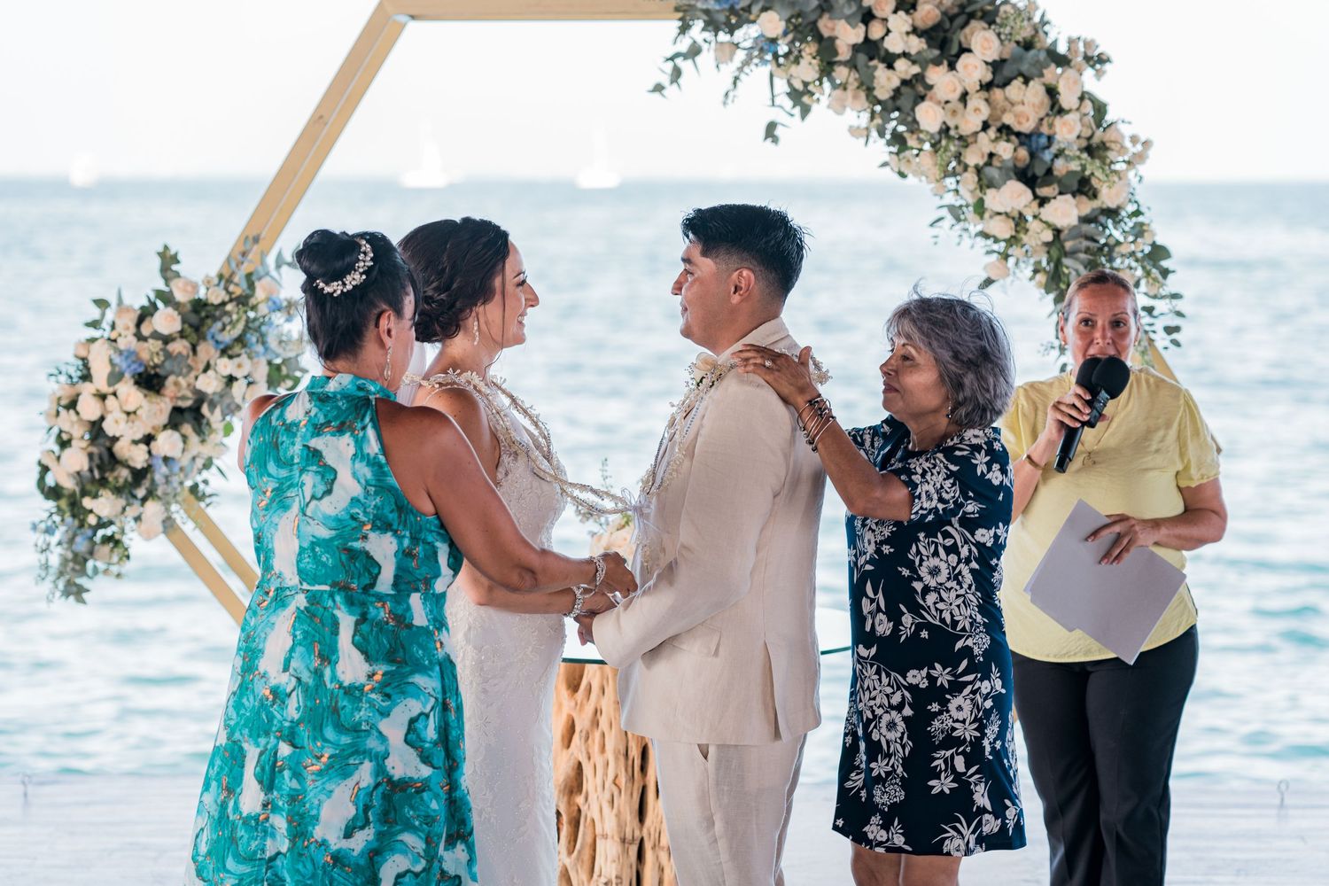 Couple exchanges vows at oceanfront hexagonal floral arch with family during beach wedding ceremony.