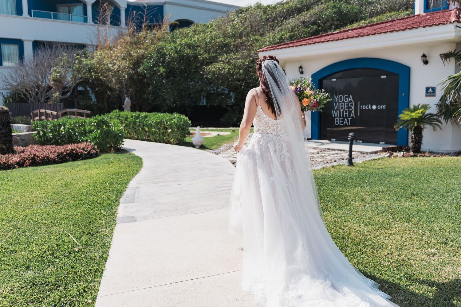 Bride in lace gown with long veil walks along garden path at coastal wedding venue.