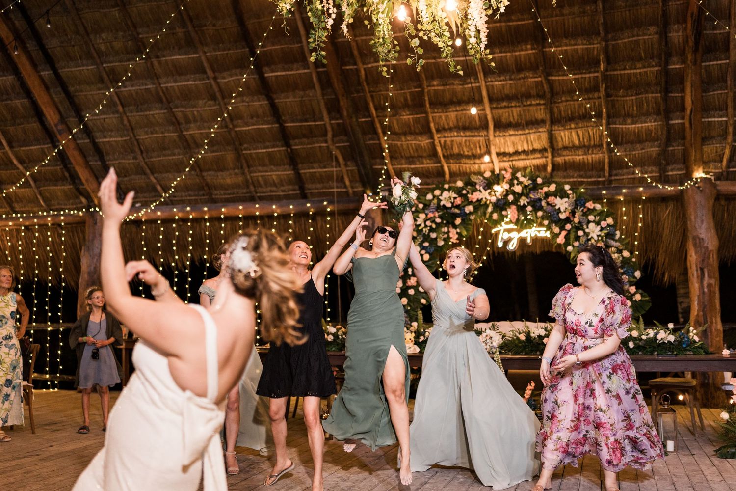 Bridesmaids jumping and celebrating during bouquet toss at decorated rustic wedding reception.