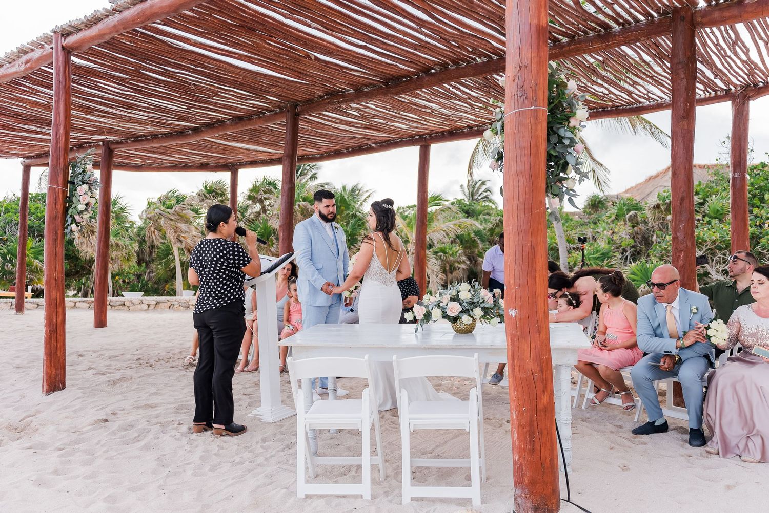 Couple exchanges vows at intimate beach wedding ceremony under rustic wooden pergola with white chairs and floral arrangements.