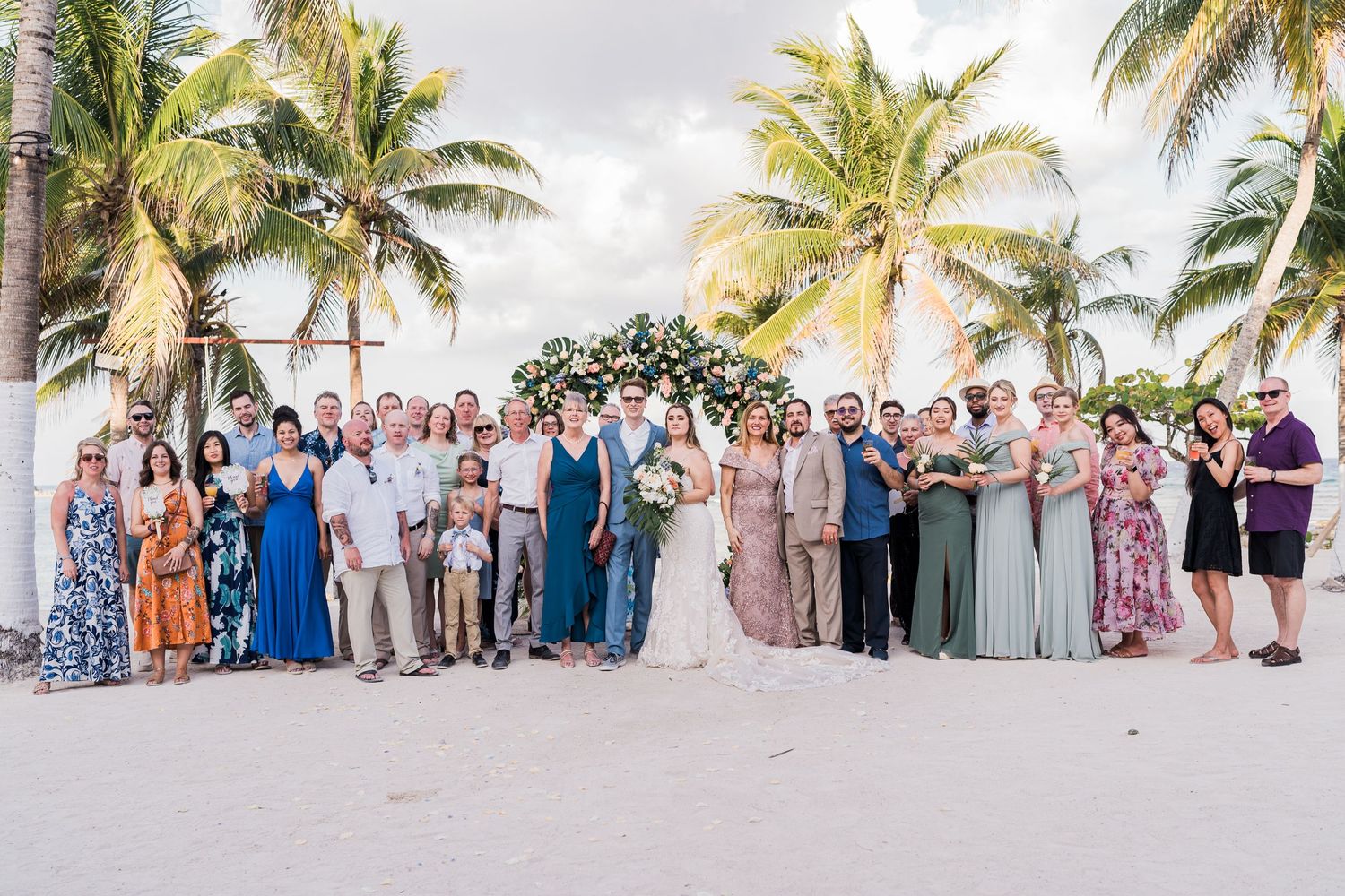 Large wedding party group portrait on sandy beach surrounded by tropical palm trees.
