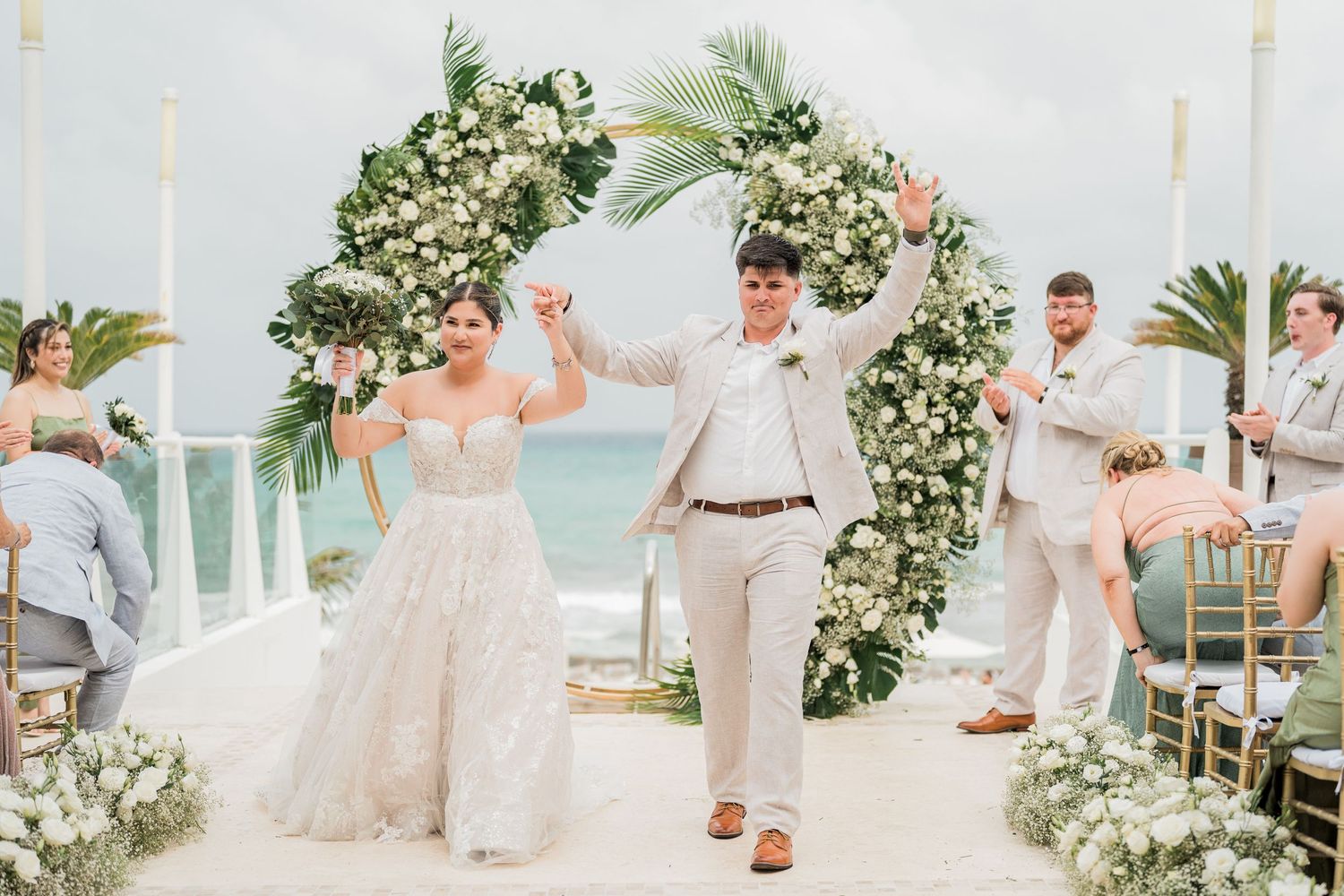 Bride and groom celebrate under floral arch at beachfront wedding ceremony with guests applauding.