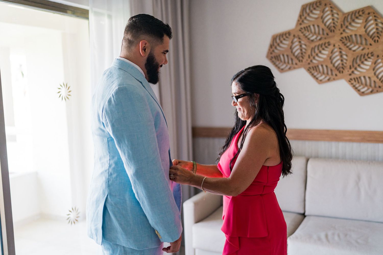Groom in light blue suit receiving a gift from a smiling woman in a red dress in a bright room.