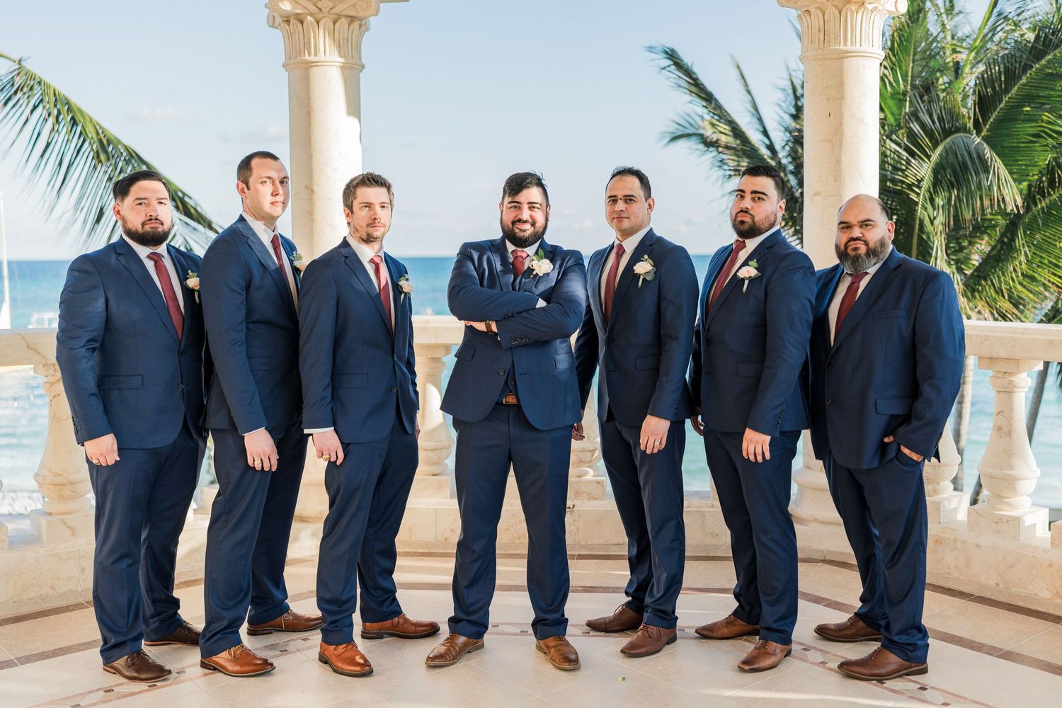 Groom in navy suit with groomsmen posing confidently between white marble columns at a tropical beachfront venue.