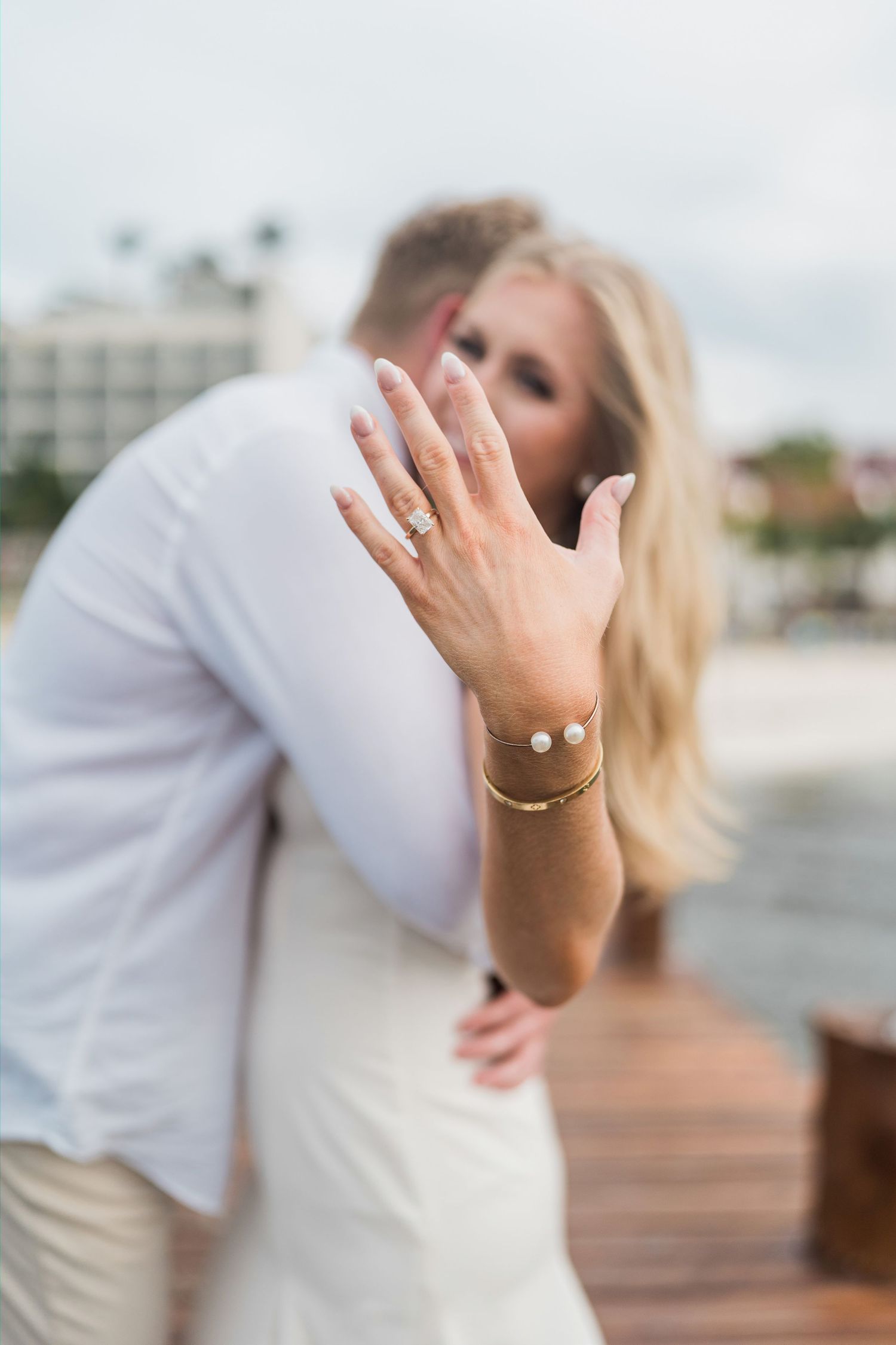Cancun Engagement Photoshoot - Blonde bride shows off engagement ring and gold bracelet while embracing groom on waterfront dock.
