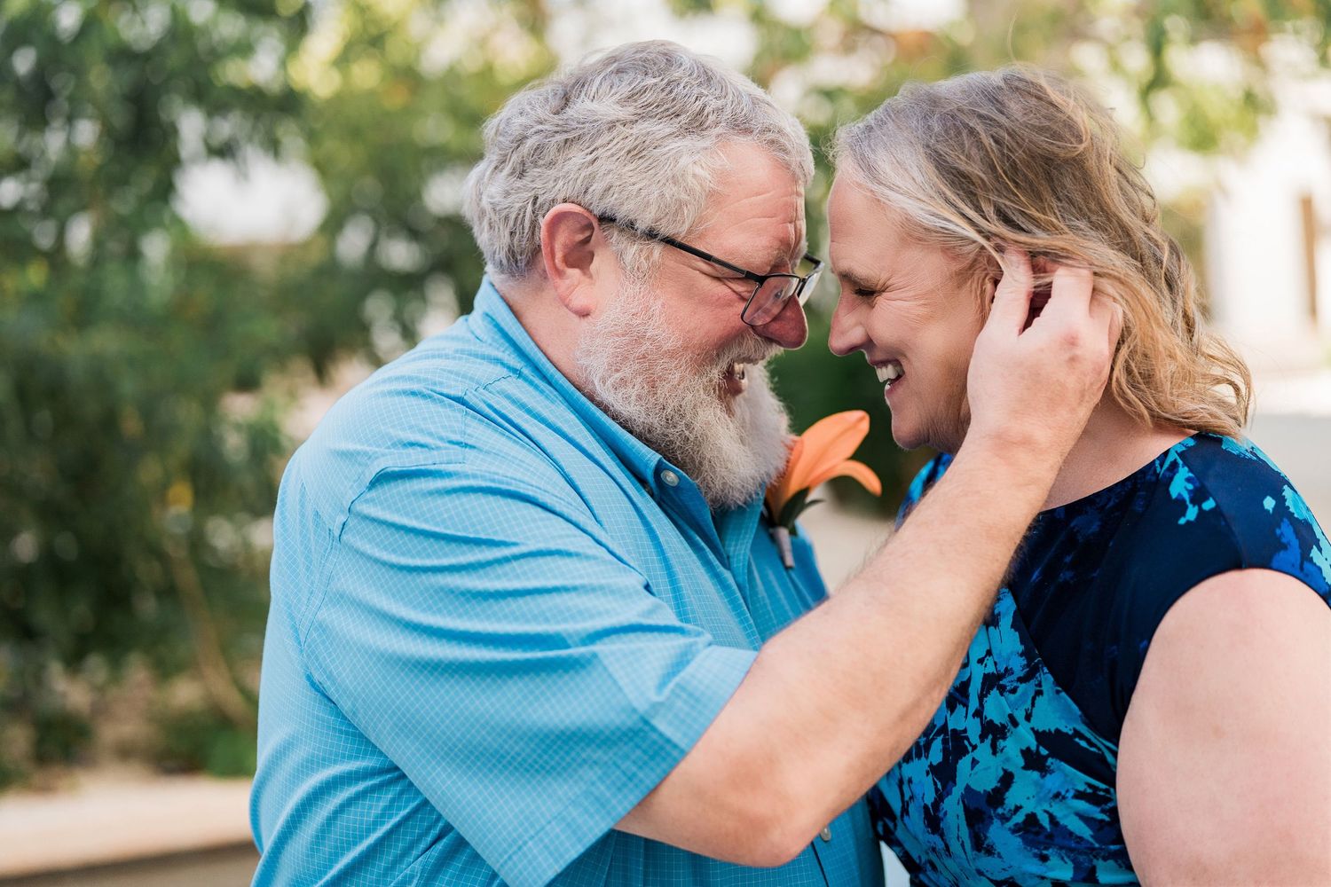 Secrets Akumal Wedding - Older couple sharing a tender moment, man in blue shirt with orange flower touching womans face, smiling outdoors.