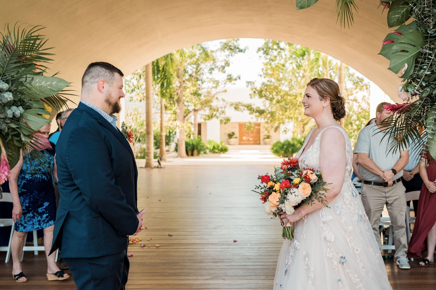 Secrets Akumal Wedding - Bride and groom exchange vows under an arch with tropical greenery at an outdoor wedding ceremony.