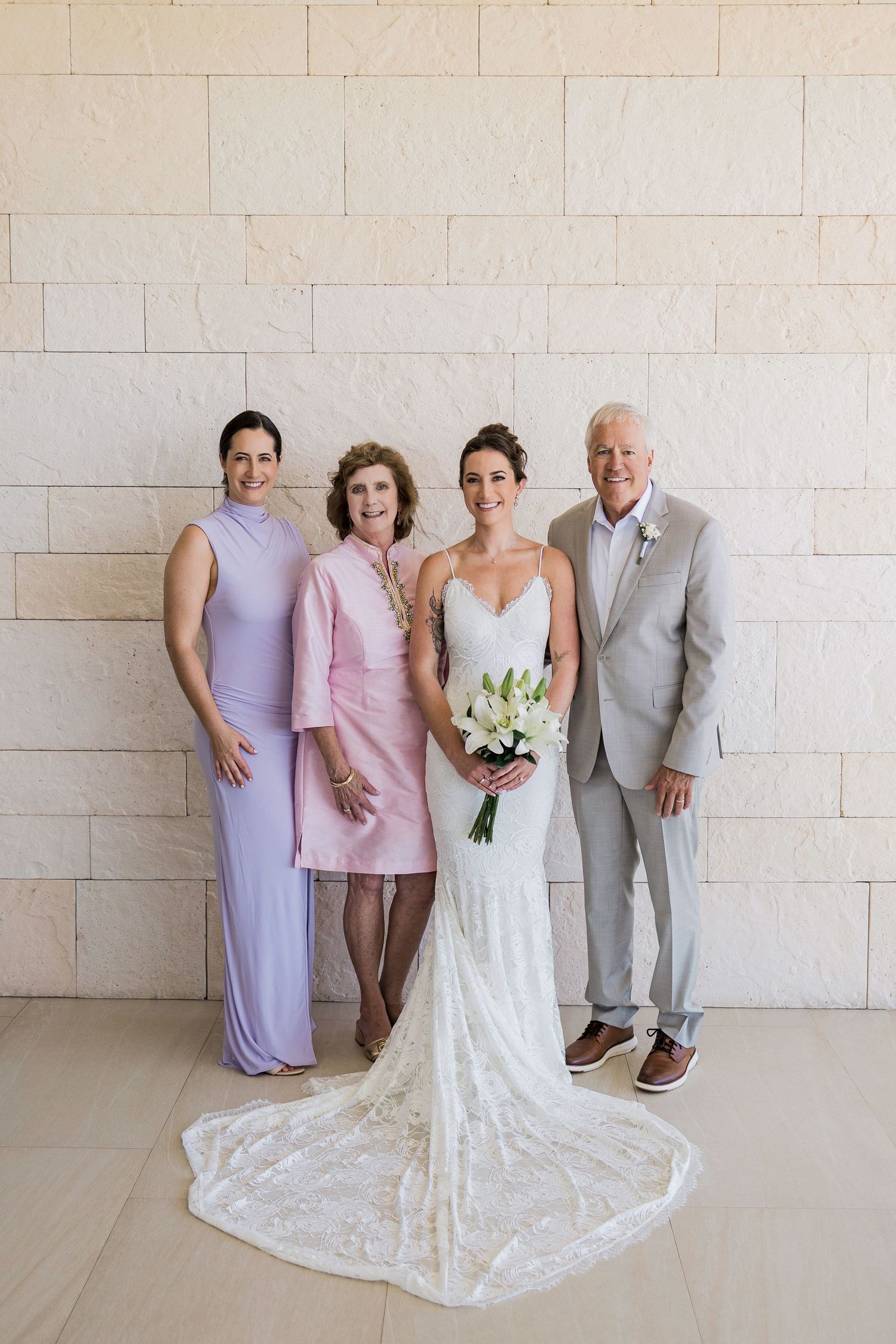 Bride in white lace gown holds bouquet, posing with family in elegant attire against a stone wall.