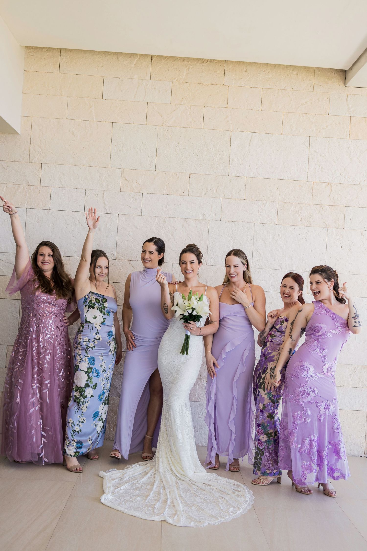 Bride in white gown poses with six bridesmaids in purple dresses, holding a white bouquet against a stone wall.