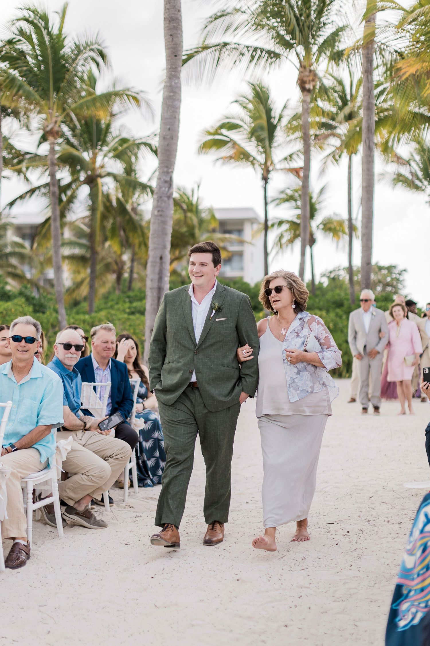 Groom in olive suit escorts woman in gray dress down sandy beach wedding aisle lined with guests and palm trees.