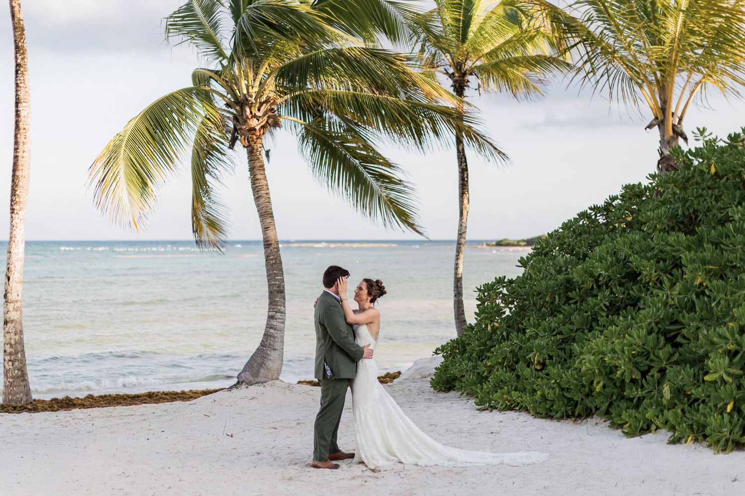 Bride and groom share a romantic moment on a tropical white sand beach with palm trees and turquoise ocean views.
