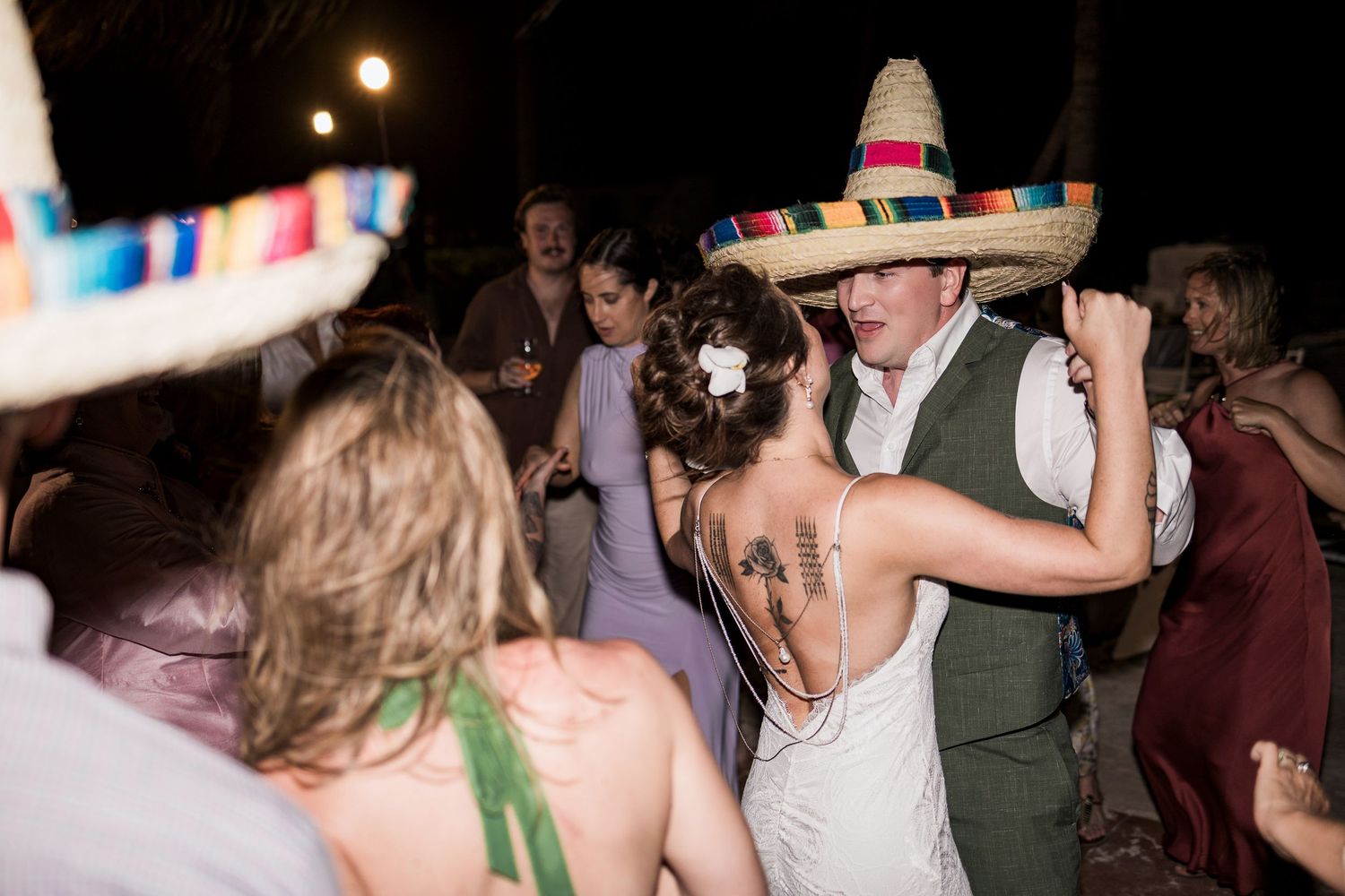 Couple dancing at wedding reception wearing colorful sombreros, bride in backless gown with tattoo, festive outdoor night celebration.
