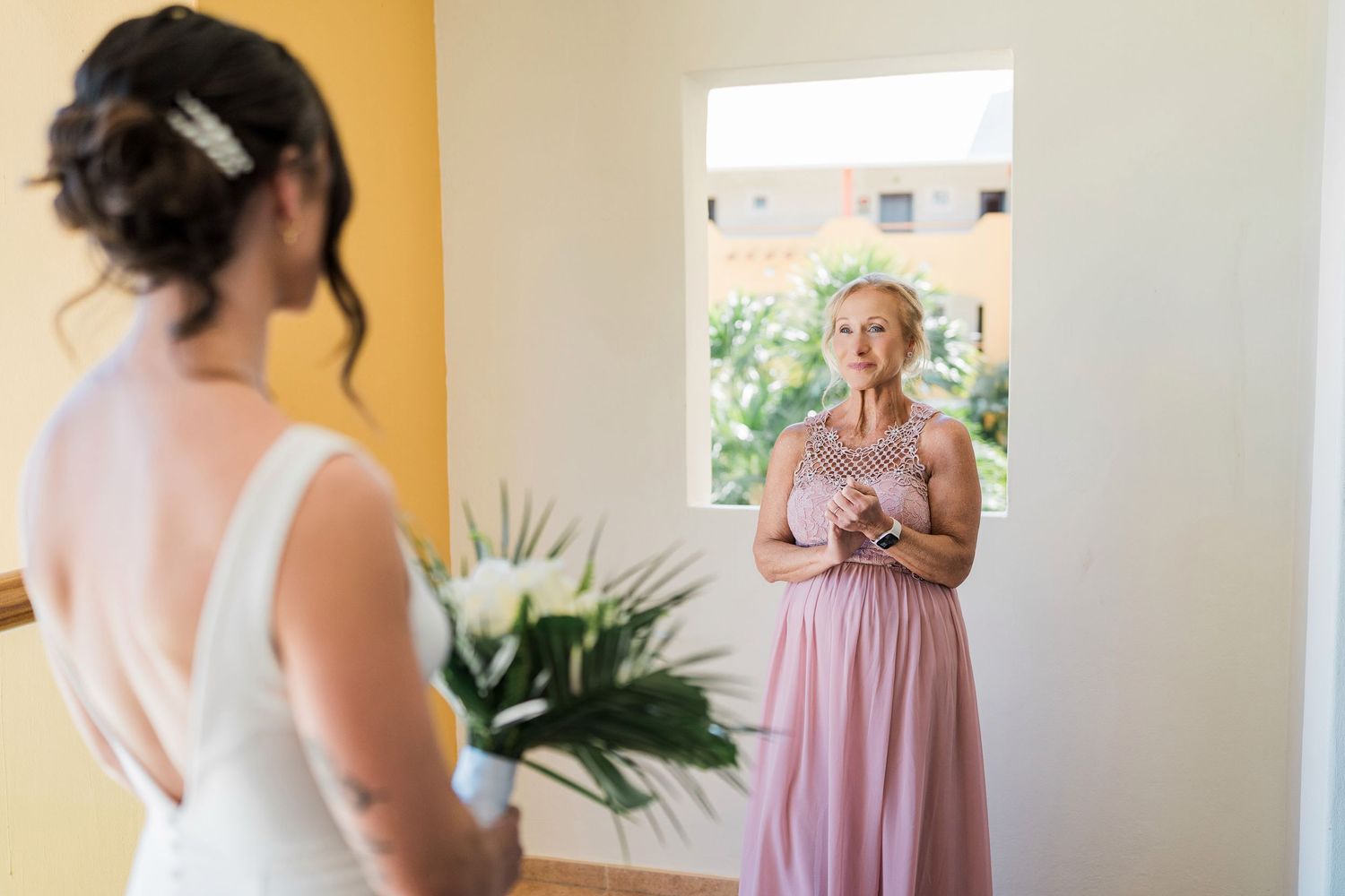 Bride in white gown holding bouquet faces her mother in pink dress during an emotional wedding morning preparation.